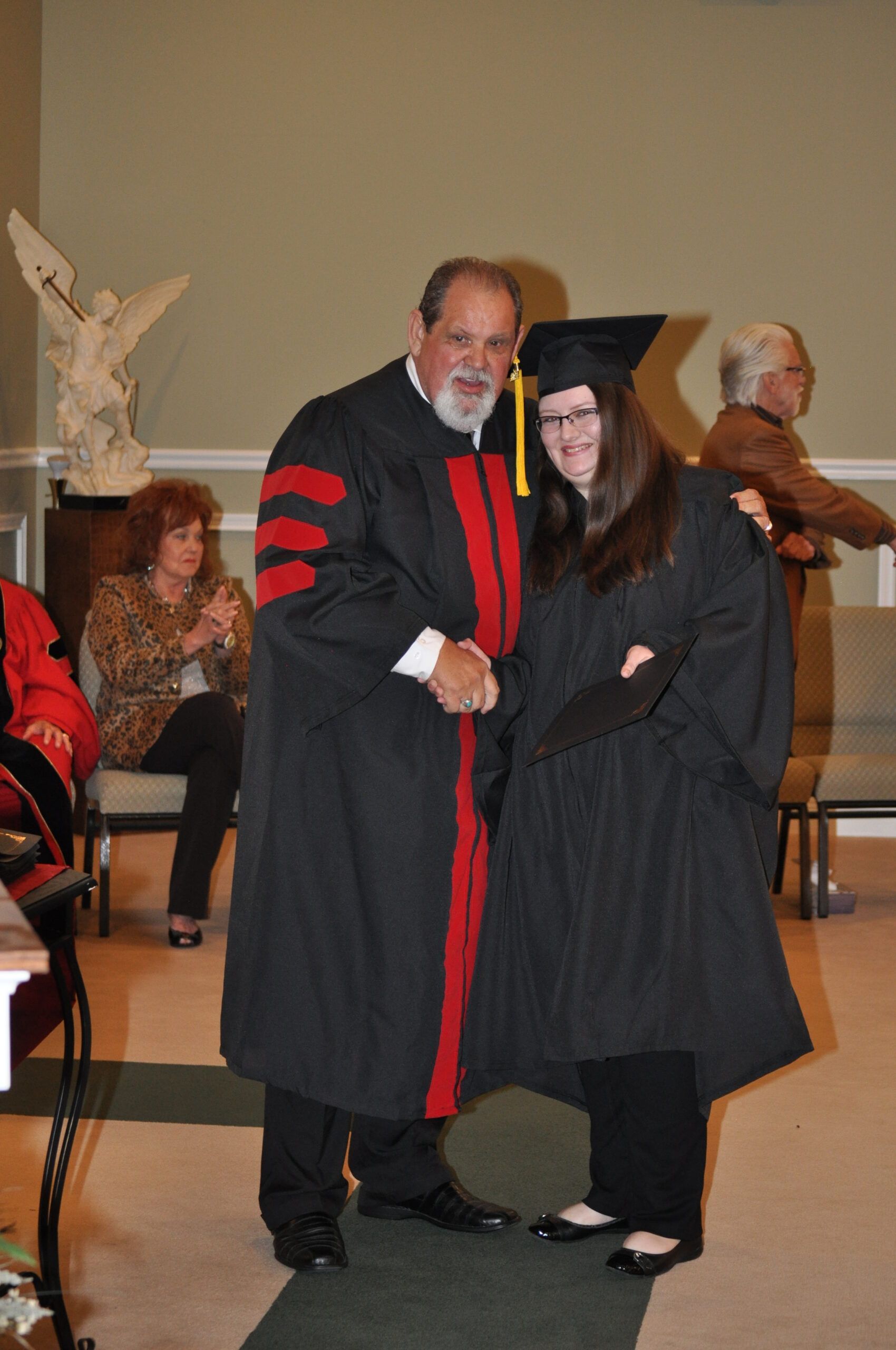 A graduate in cap and gown shakes hands with a person in academic regalia; others watch.