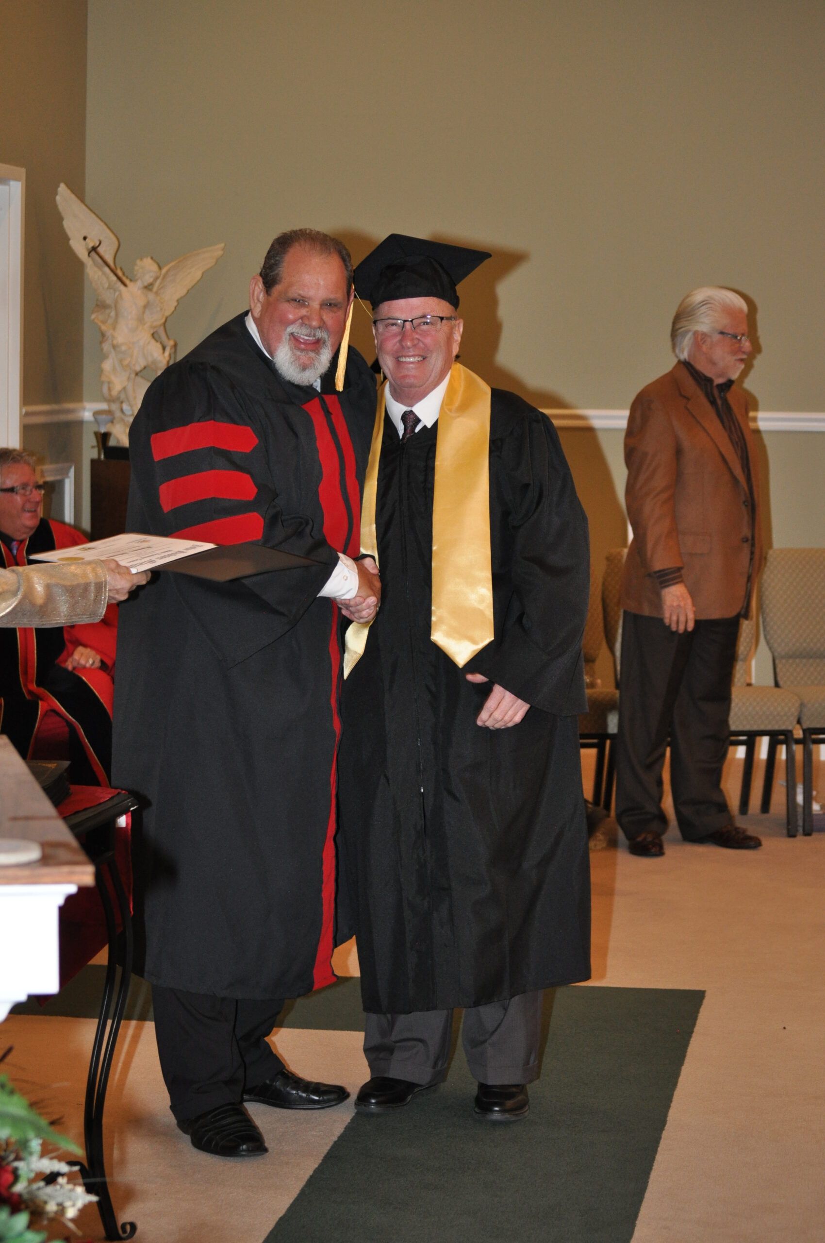 Man in graduation gown receiving diploma from another, in a hall.