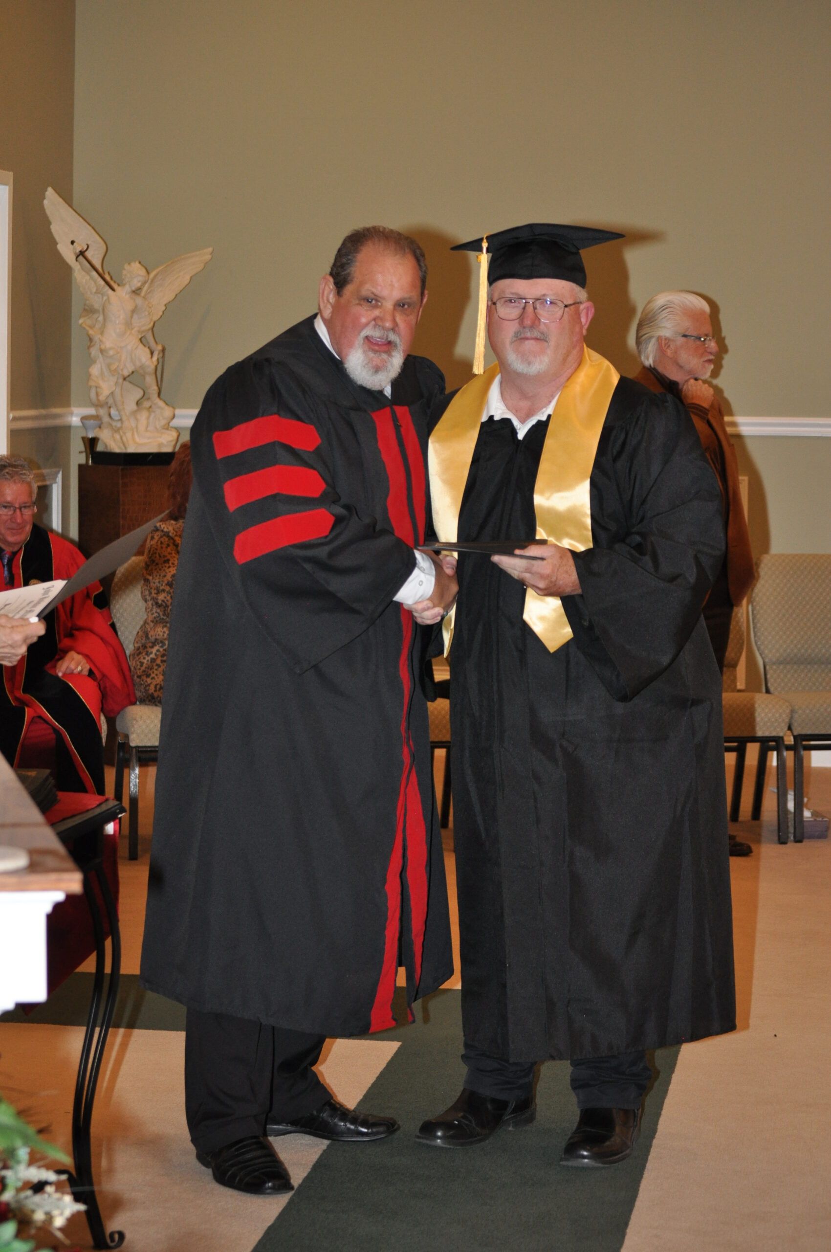 Man in graduation cap and gown receives diploma from robed man indoors.