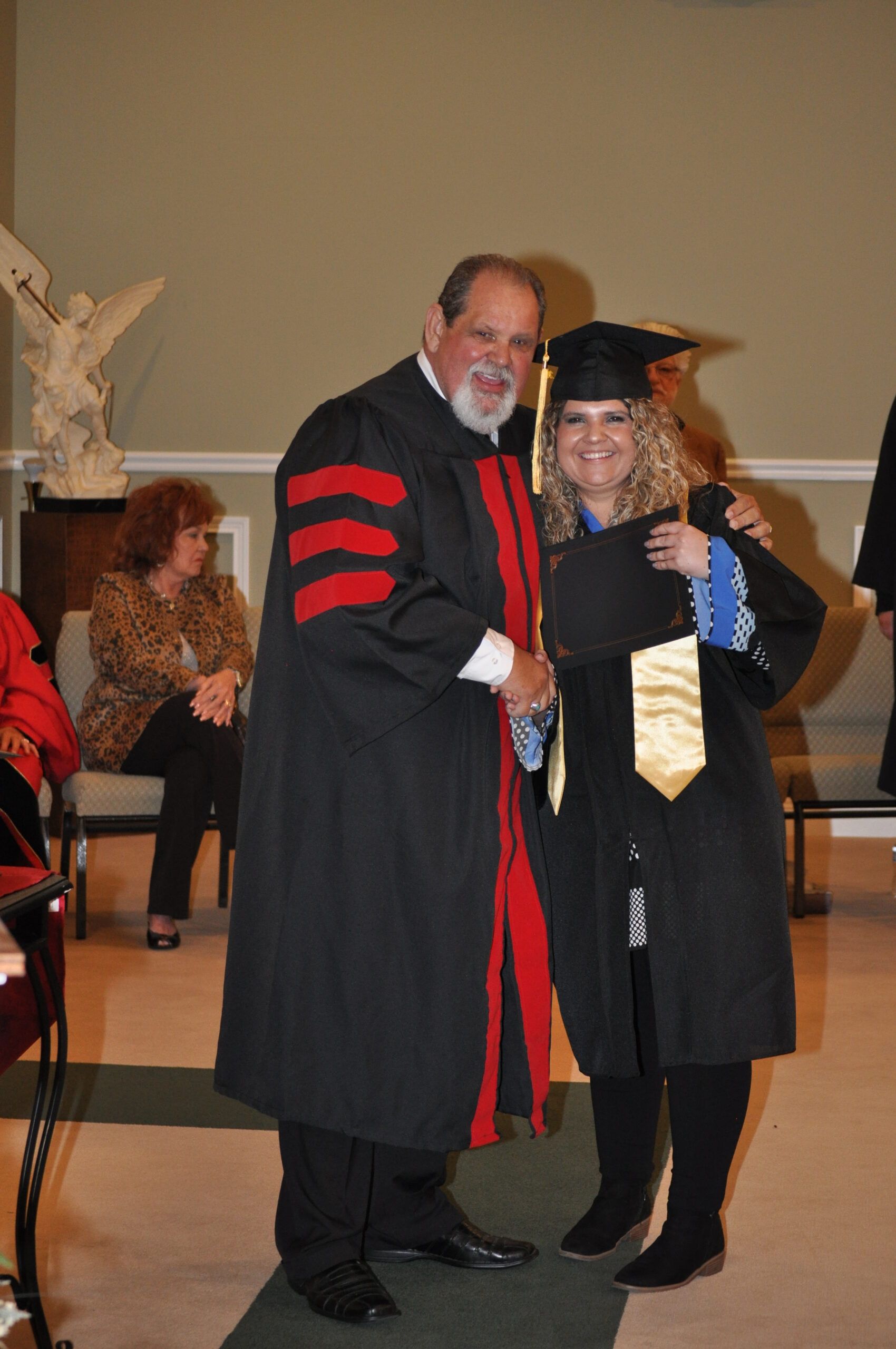 Man in academic robe congratulates a graduate wearing cap and gown; they are smiling.