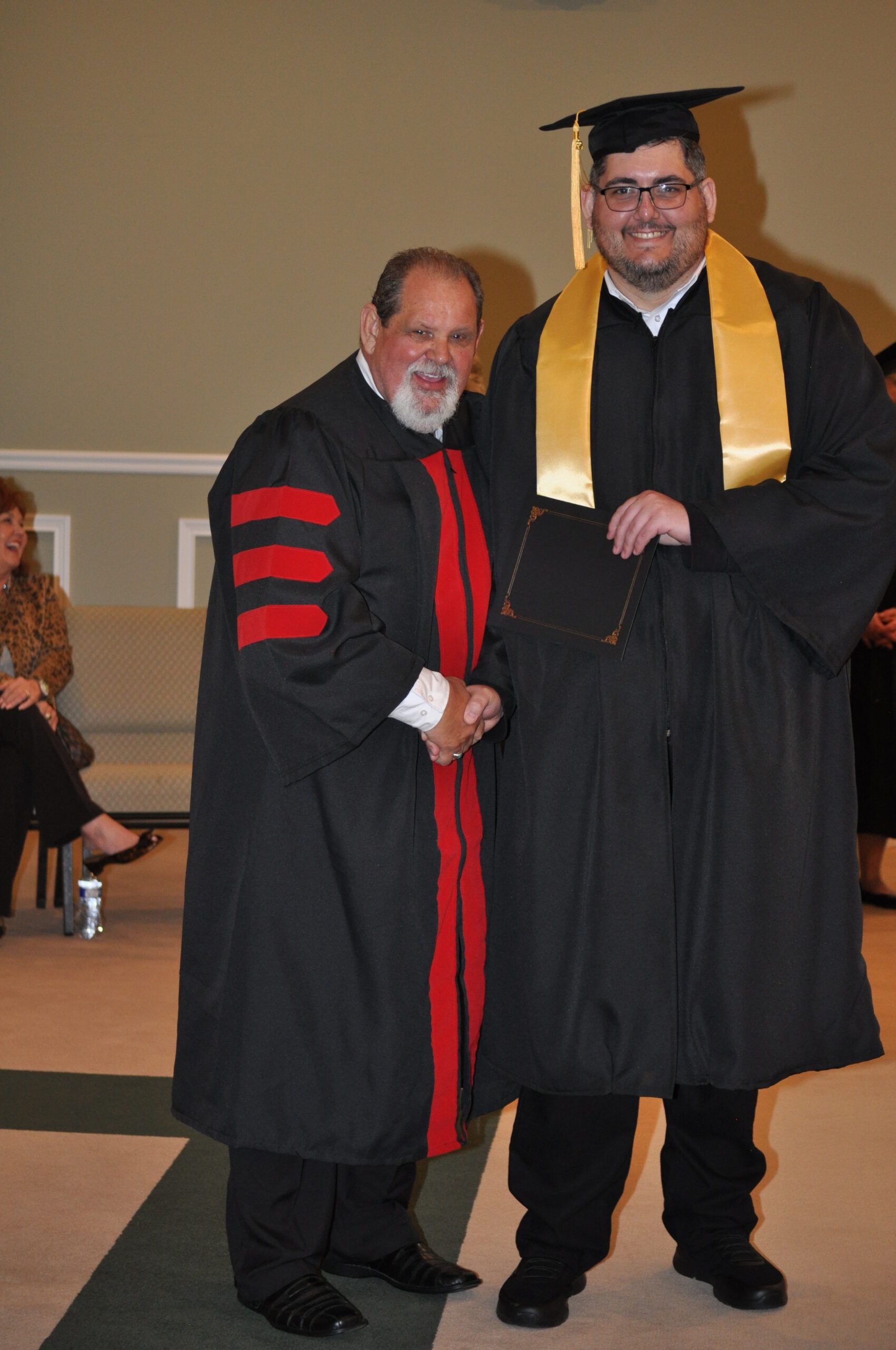 Two men in graduation attire shake hands on a stage. One wears a black robe with red accents, the other a black robe with a gold sash and cap.