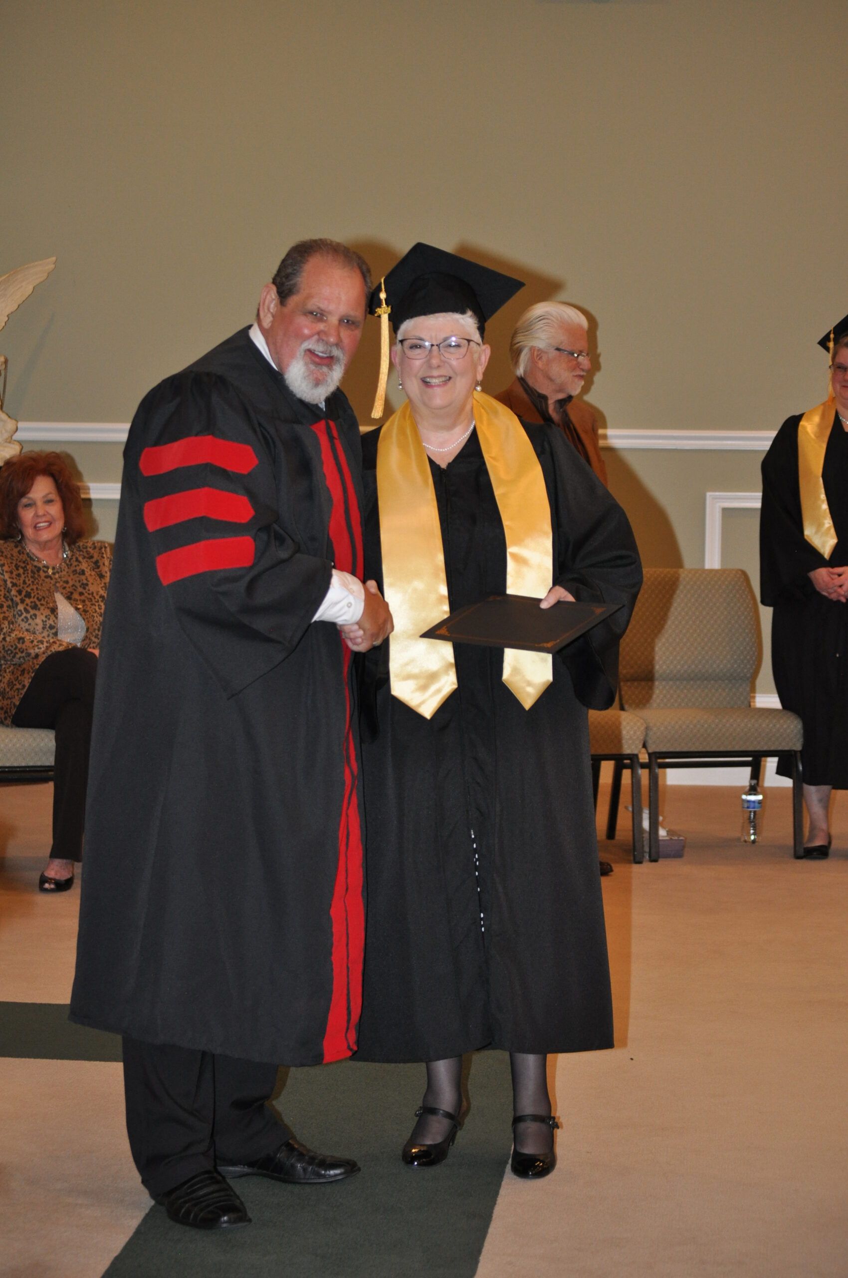 Man in academic robe shakes hands with graduate wearing cap and gown.