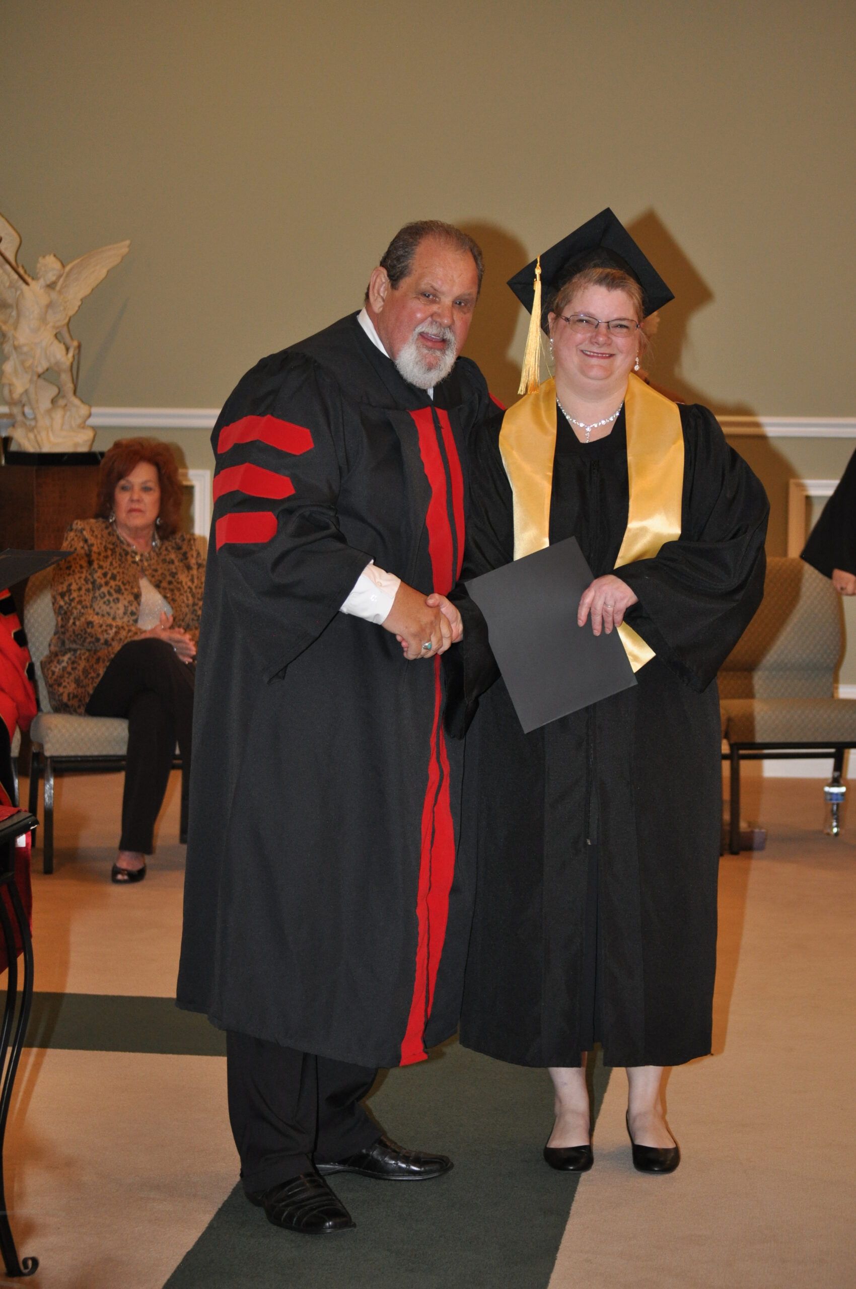 Man in academic robes shakes hands with a person in graduation attire, holding a diploma.
