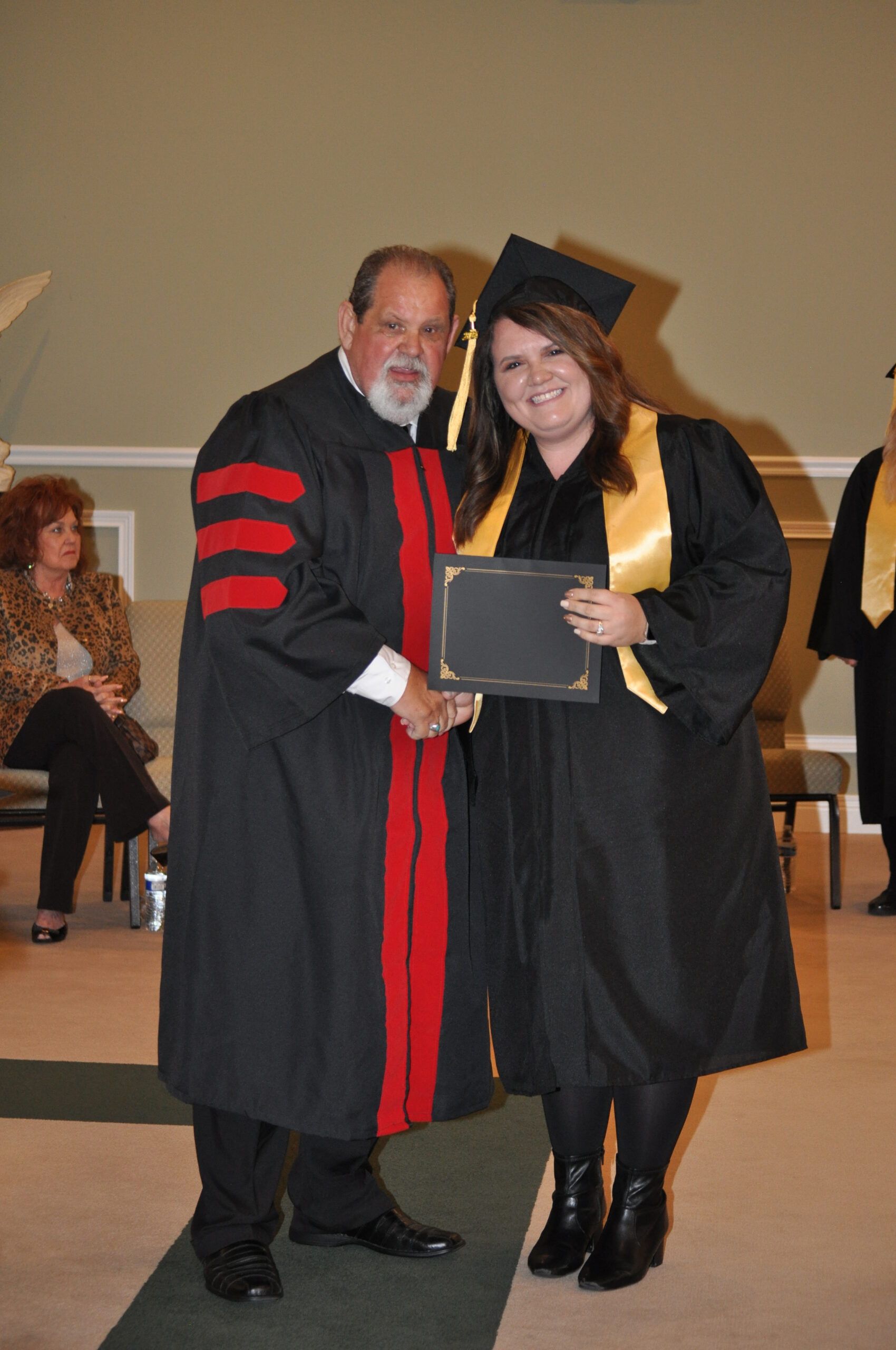Man in graduation robe handing diploma to graduate in cap and gown. Inside a building.