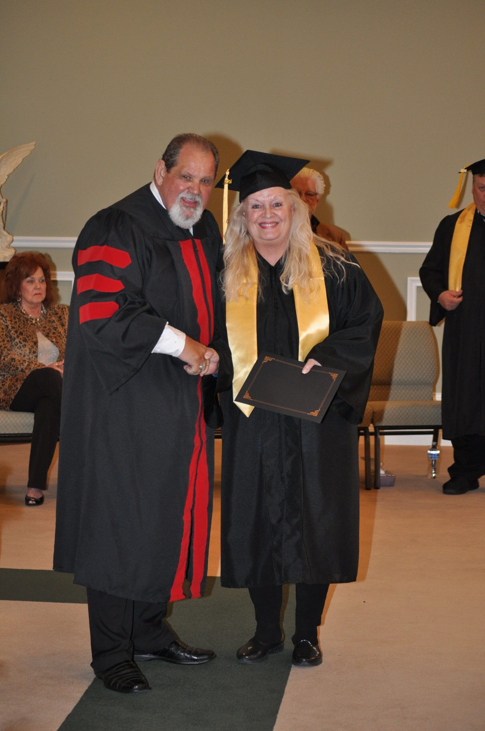 Graduation ceremony: a person in cap and gown receives a diploma from a robed individual; cream-colored interior.