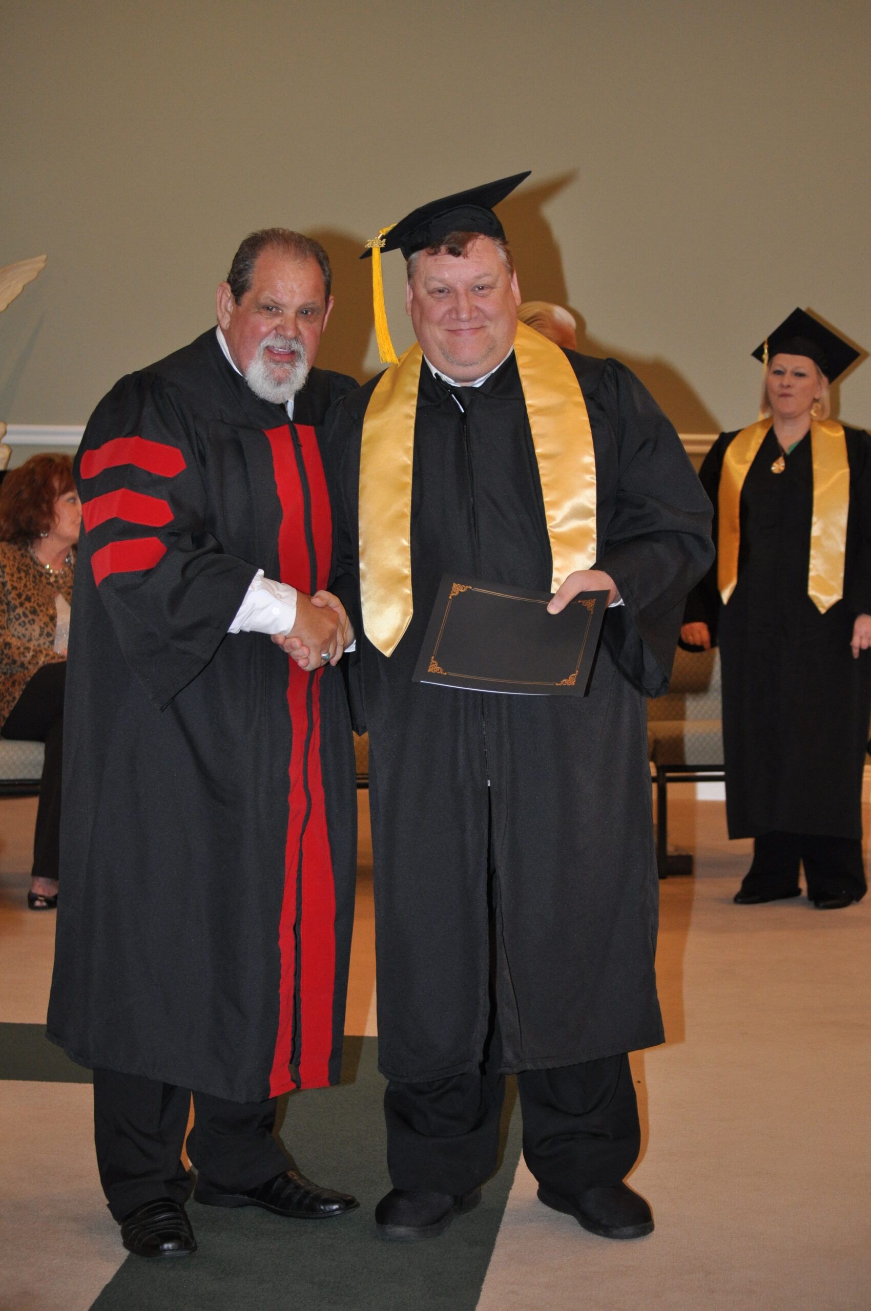 Man in graduation gown shakes hands with a man in academic regalia, inside a building.