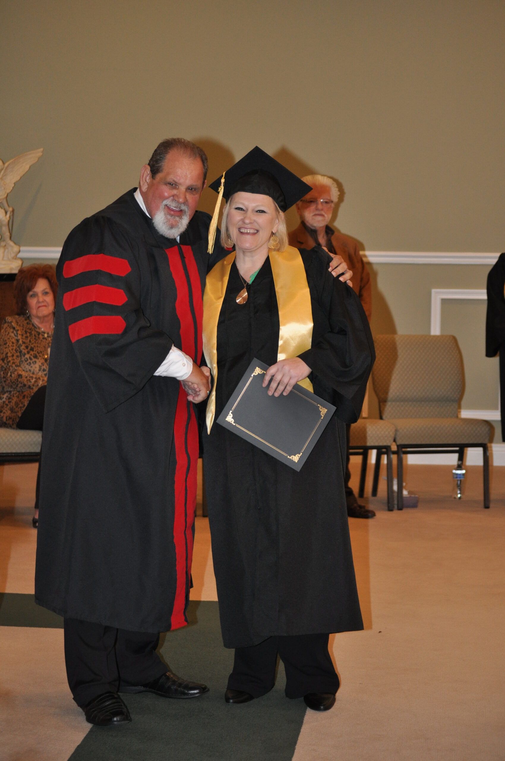 Person in graduation regalia shakes hands with another, holding a diploma in a church setting.