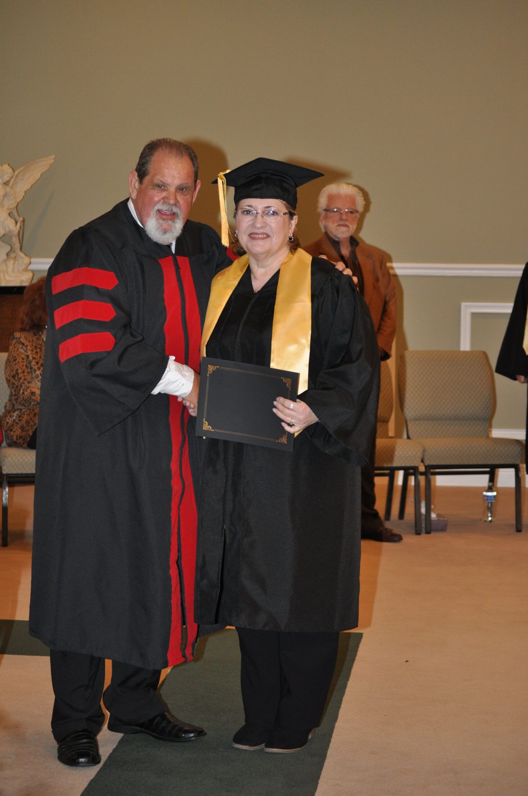Man in academic robe shakes hands with graduate wearing cap and gown; another person stands nearby in an auditorium.