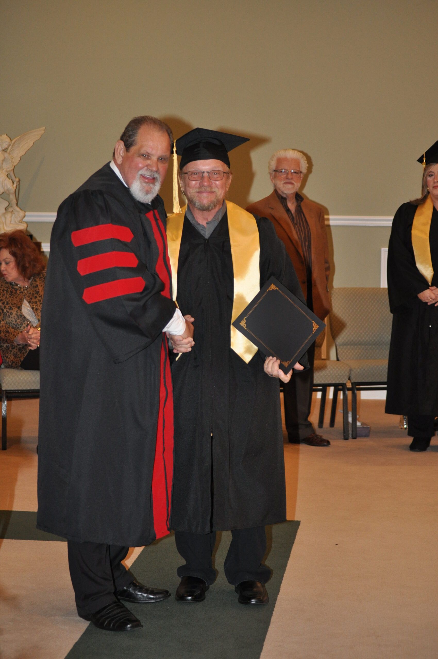 Man in graduation gown shakes hands with another man in academic regalia.