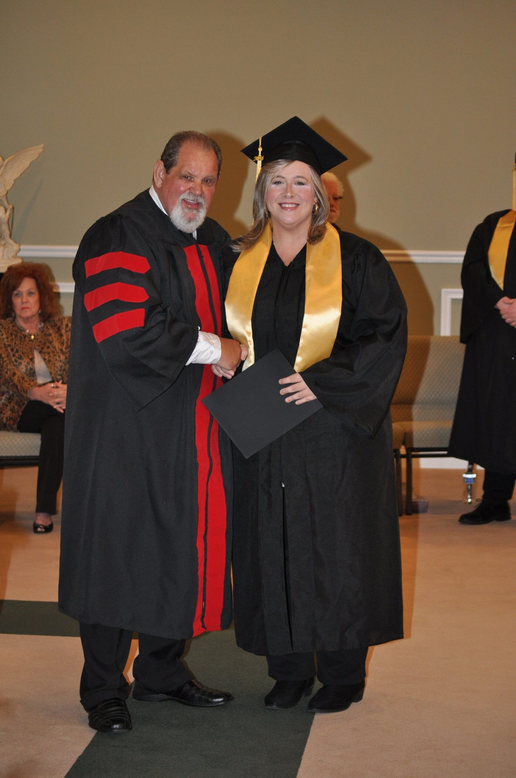 Man in academic robe shakes hands with a woman in a graduation gown, certificate in hand. Indoor ceremony.