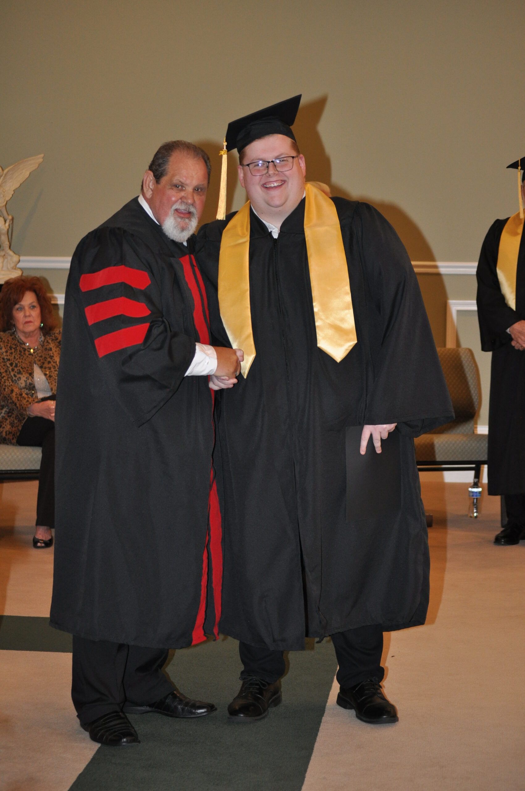 Man in cap and gown shakes hands with a person in academic regalia, in a graduation setting.
