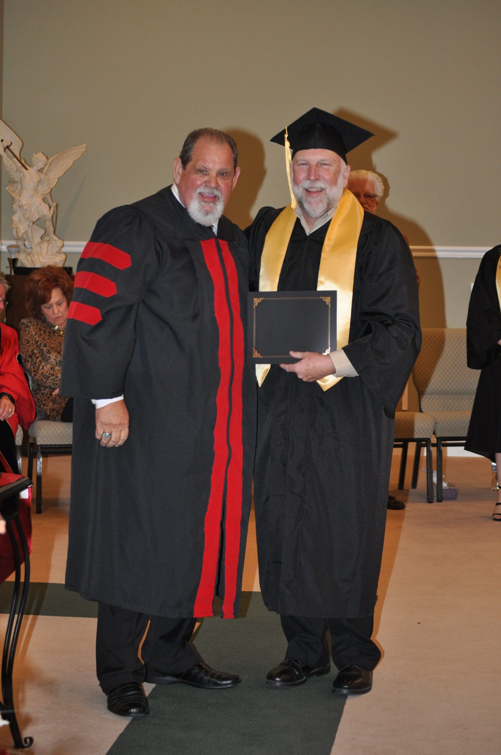 Two men in graduation attire pose indoors. One wears a black robe with red accents and the other a black gown, cap, and gold stole holding a diploma.
