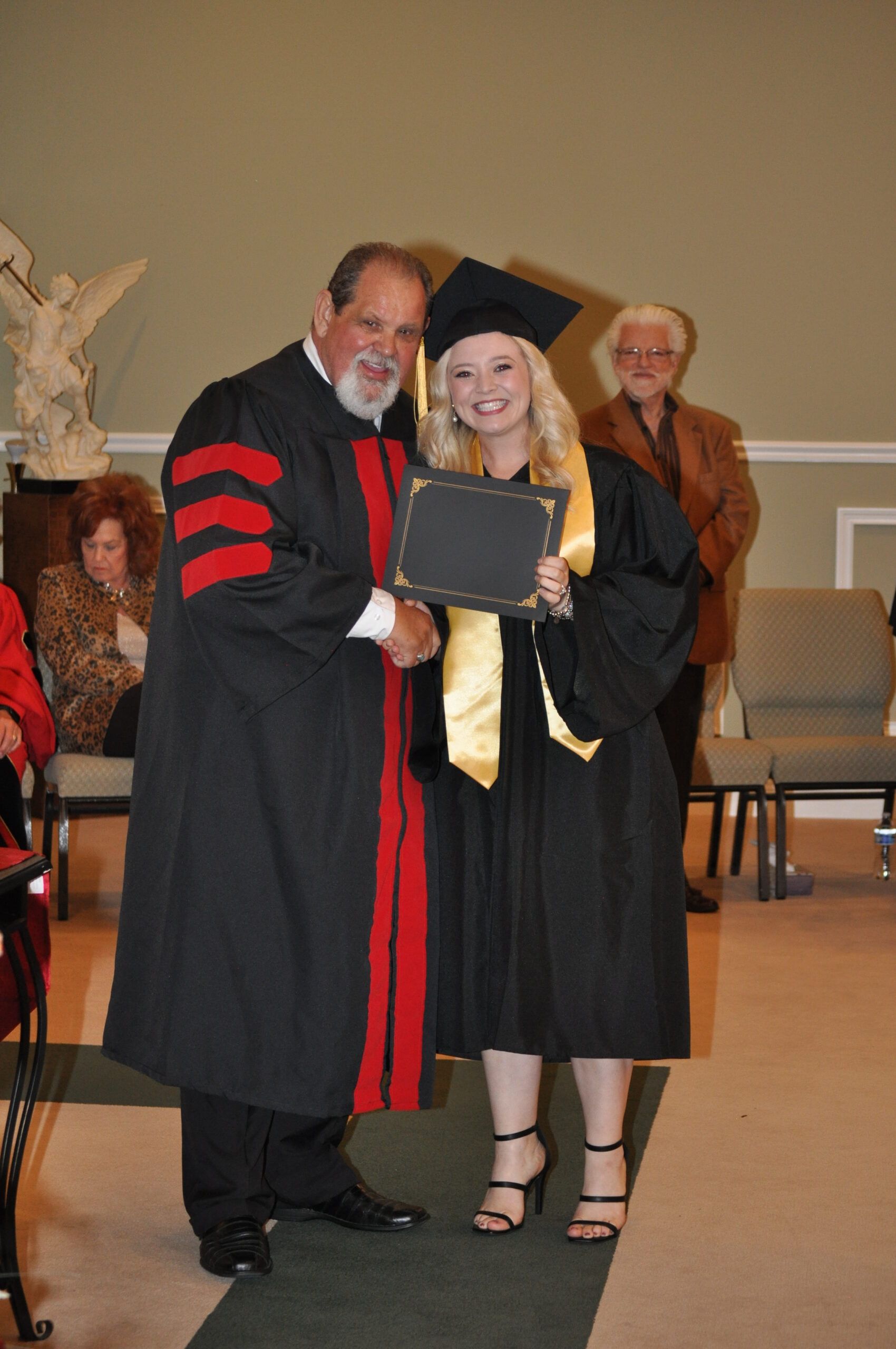 Graduation ceremony: Man in academic robe shaking hands with a woman in cap and gown, holding a diploma.
