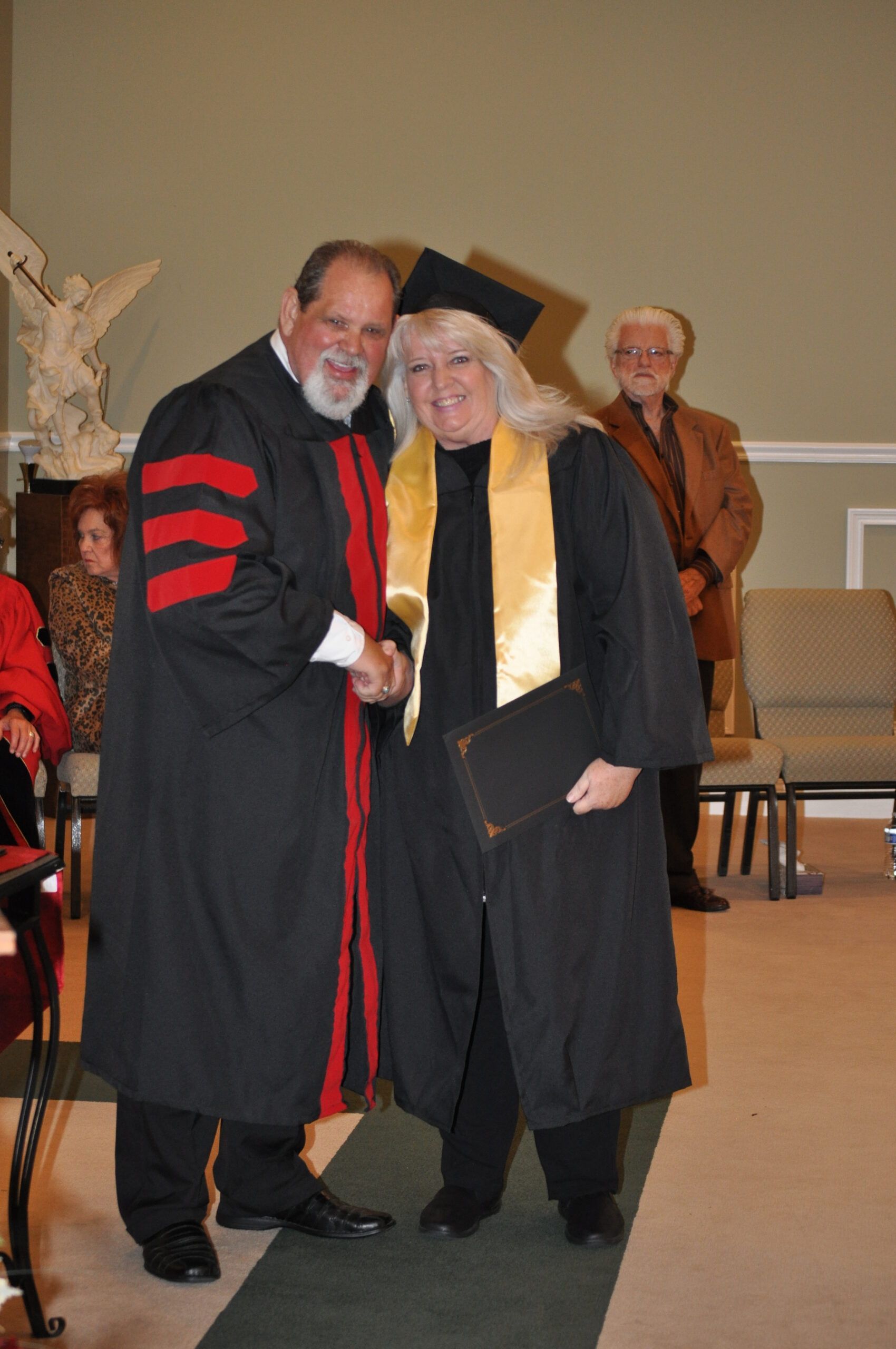 Man in academic robe shakes hands with a woman in graduation attire, both smiling.