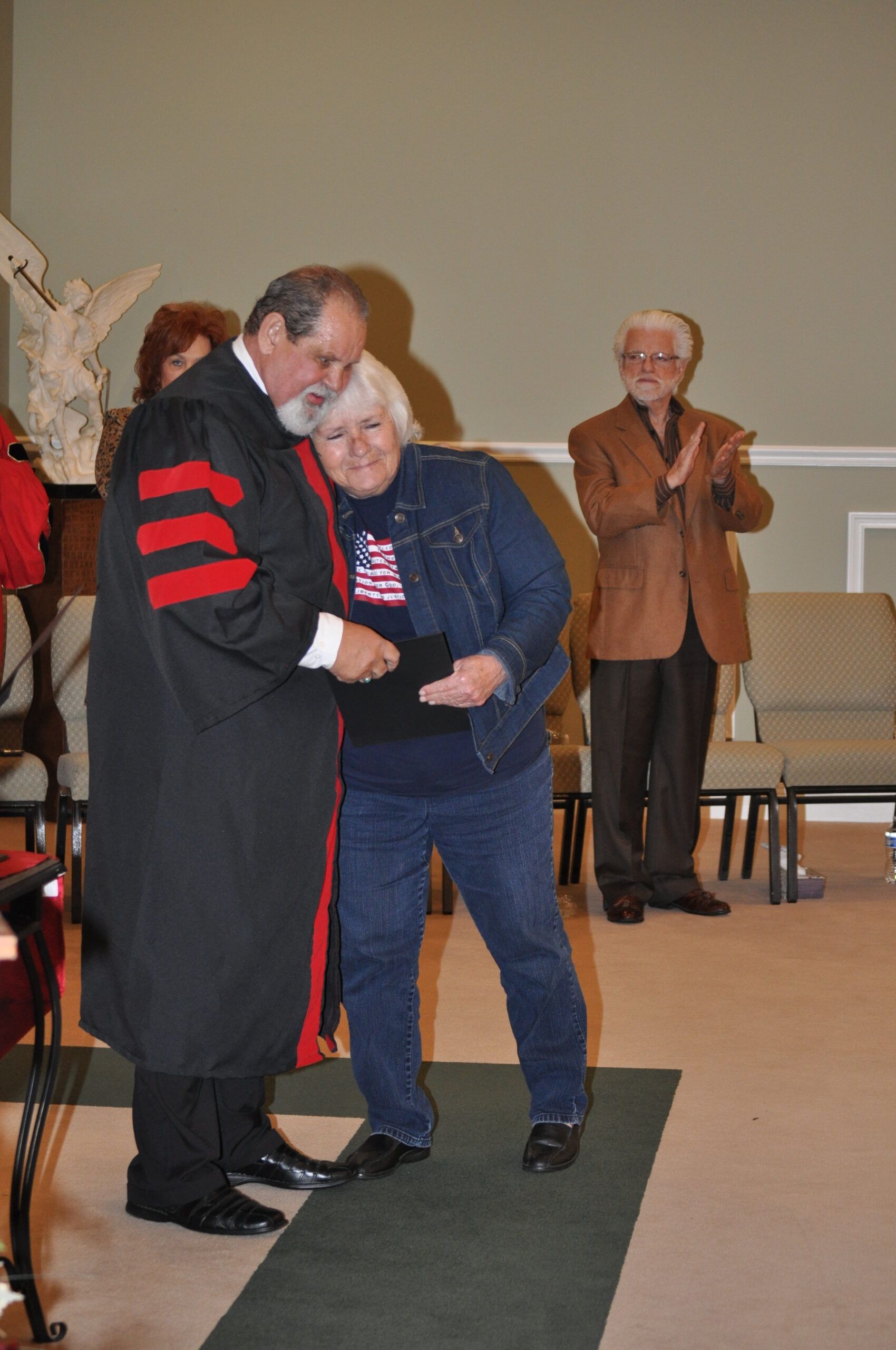 Man in academic robe presents a certificate to a woman in a denim jacket on a stage; man applauds in the background.