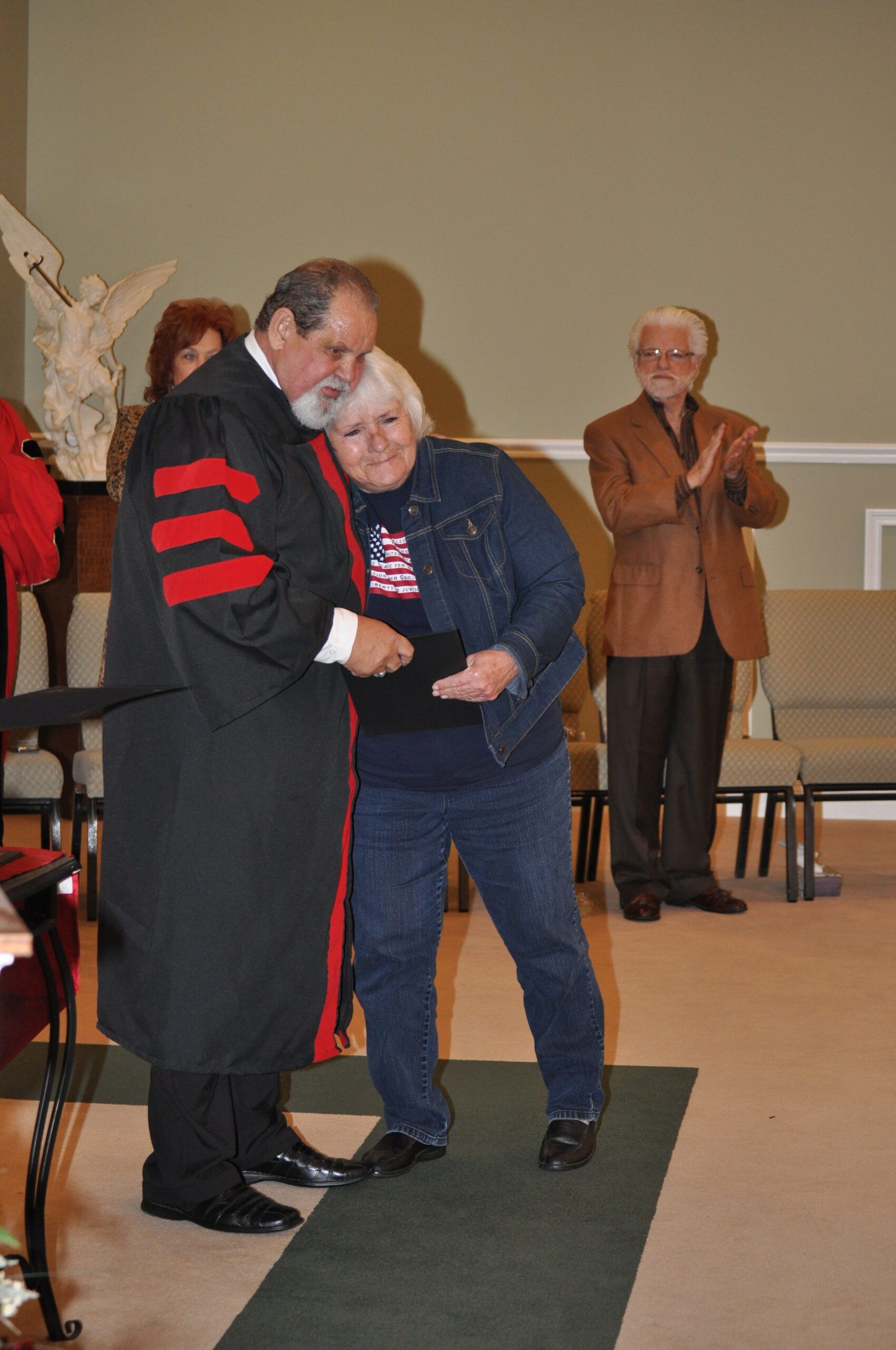 Man in academic robe hugs a woman holding a document, another man claps in a ceremony setting.