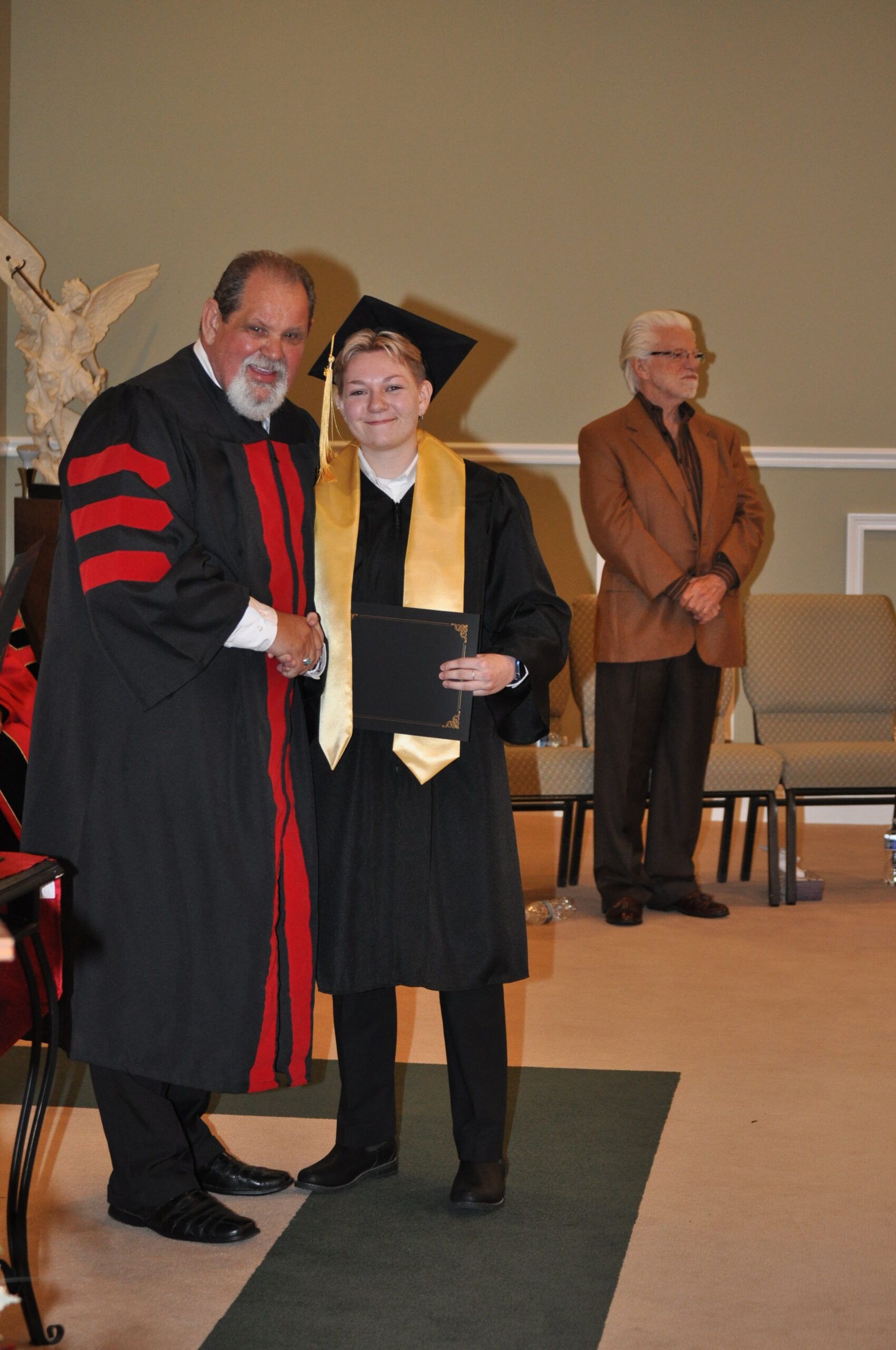 Person in graduation attire shakes hands with robed figure. Ceremony setting, other person watches.