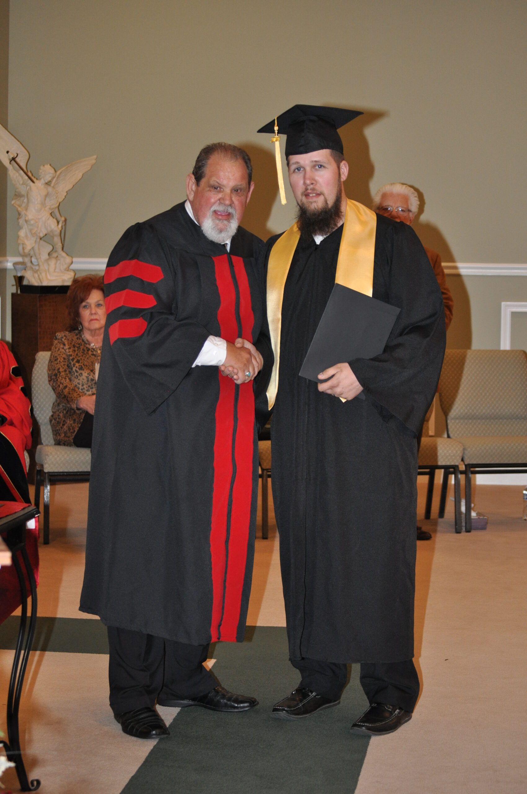 Man in graduation gown receiving diploma from a man in academic robes, indoors.