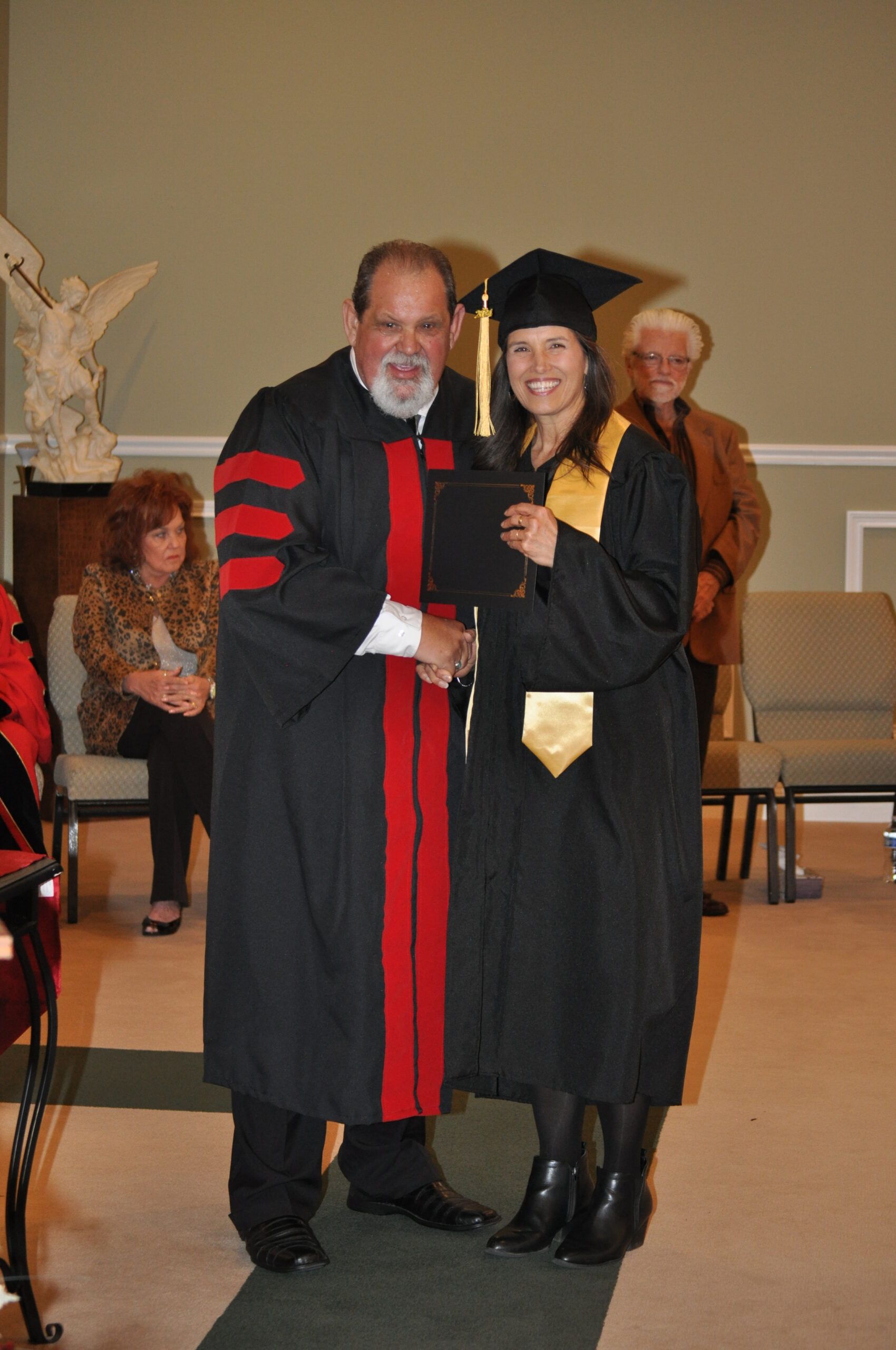 Person in graduation gown receives diploma from robed person, seated audience.
