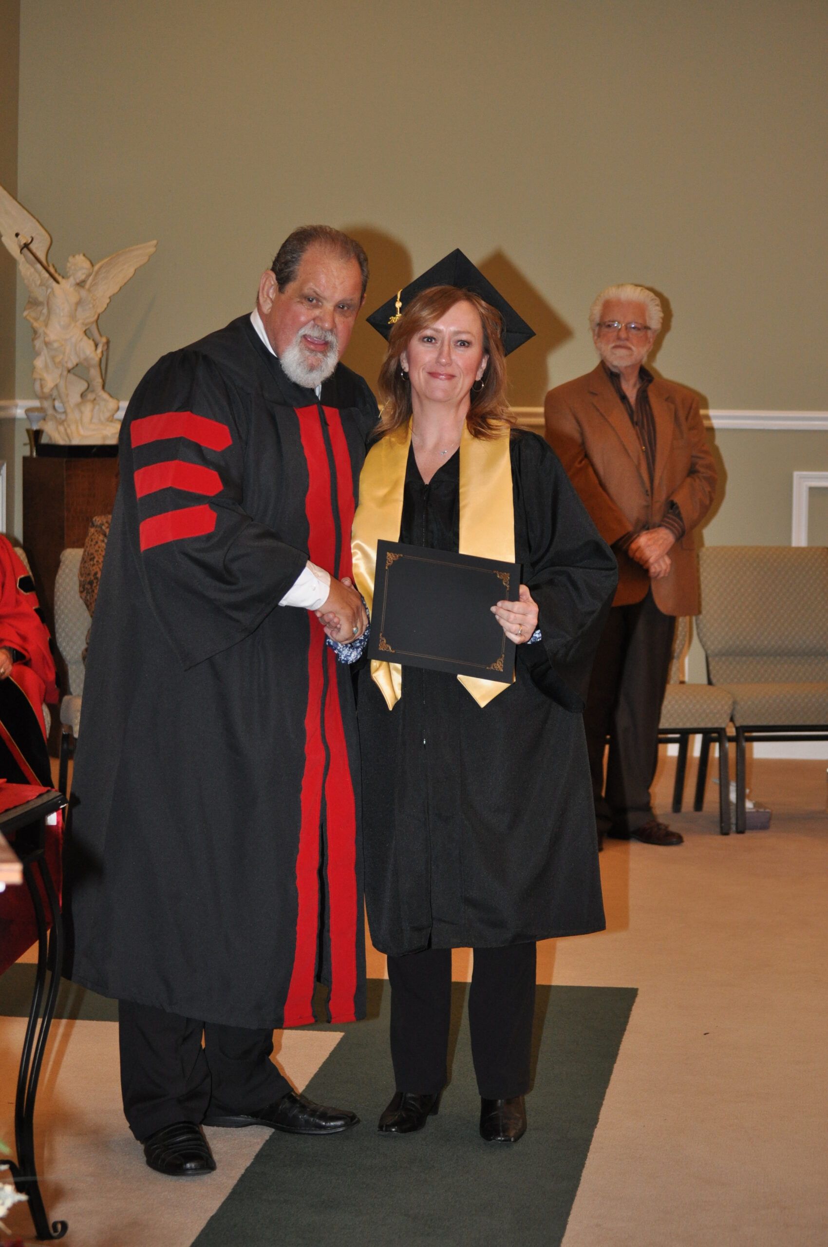 Graduation ceremony: a person in cap and gown shakes hands with a person in academic regalia, inside a building.