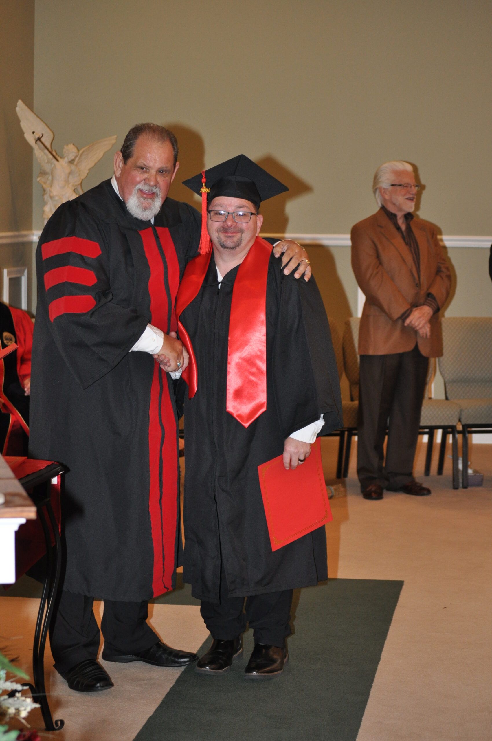 Man in graduation gown receives diploma, shaking hands. Another man with a blazer watches. Church interior.