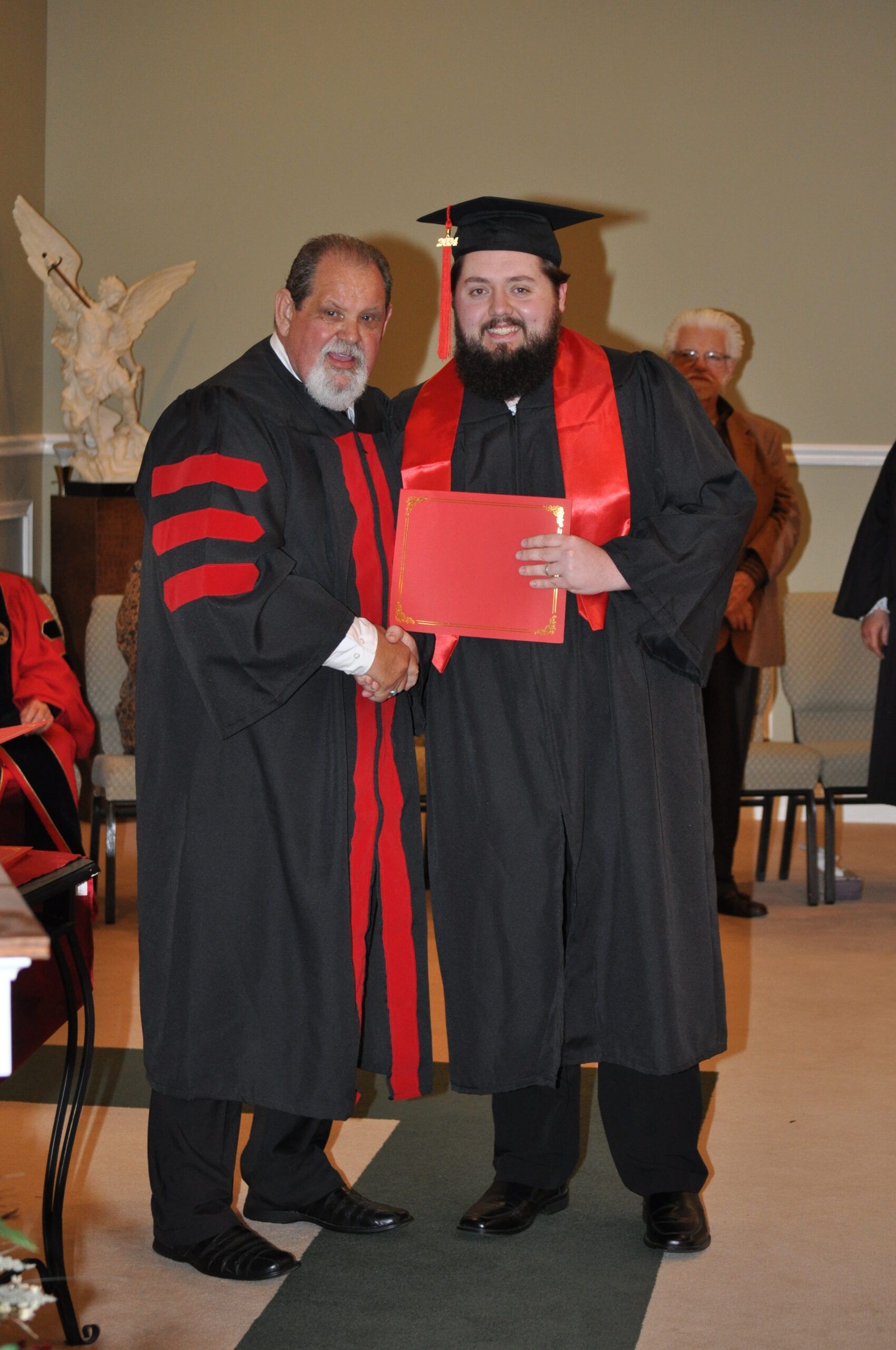 Man in graduation gown receives diploma, shakes hands with official in academic regalia, indoor setting.