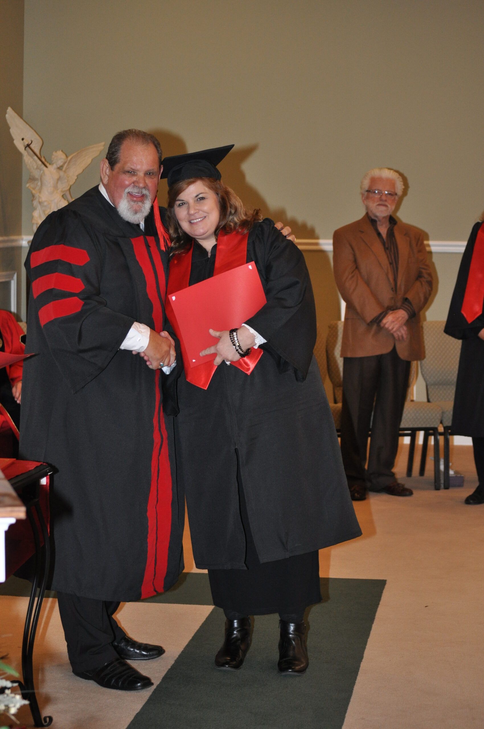 A graduate in cap and gown shakes hands with a person in academic regalia; red trim. Ceremony setting.