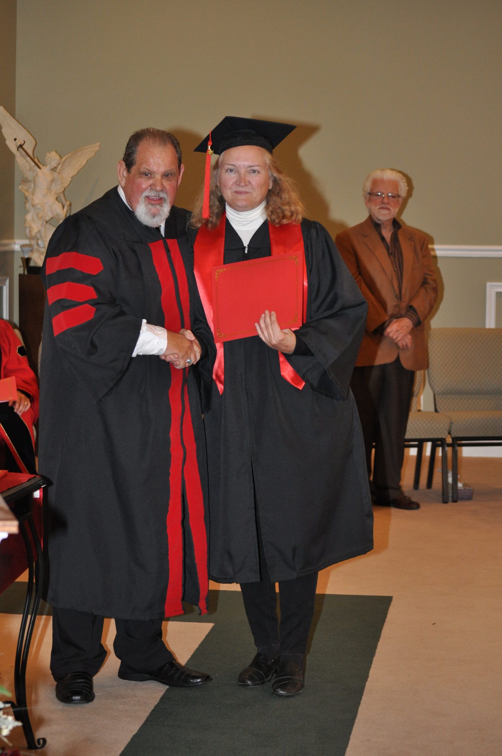 A person in graduation attire receives a diploma from a robed figure. Another man watches.