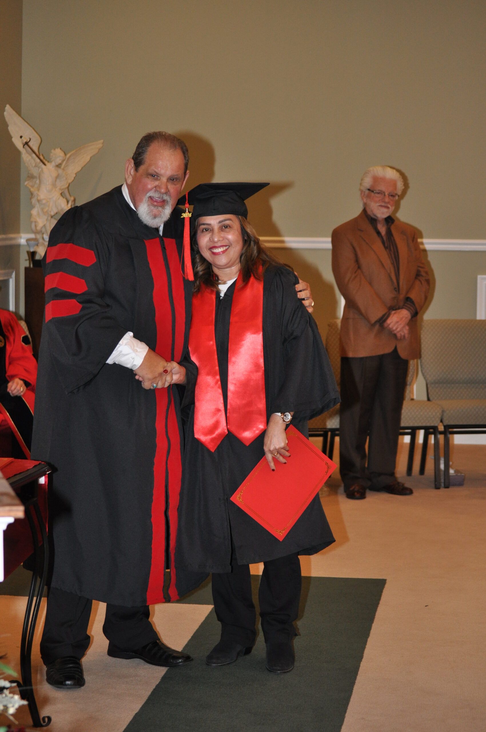 Man in academic robe congratulates woman in cap and gown, holding a diploma, inside a building.