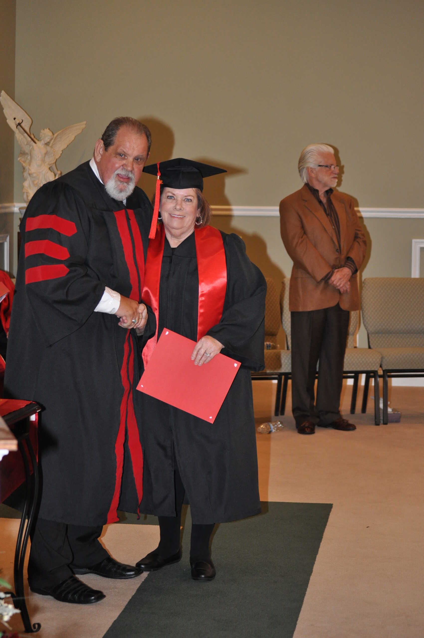 Man in academic robes shakes hands with woman in cap and gown, holding red folder. Another man observes in the background.