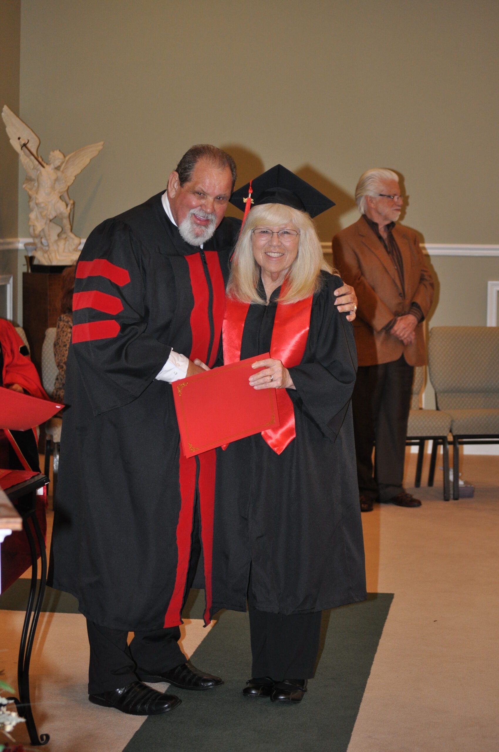 A graduate receives a diploma from a robed person indoors with another person in the background.
