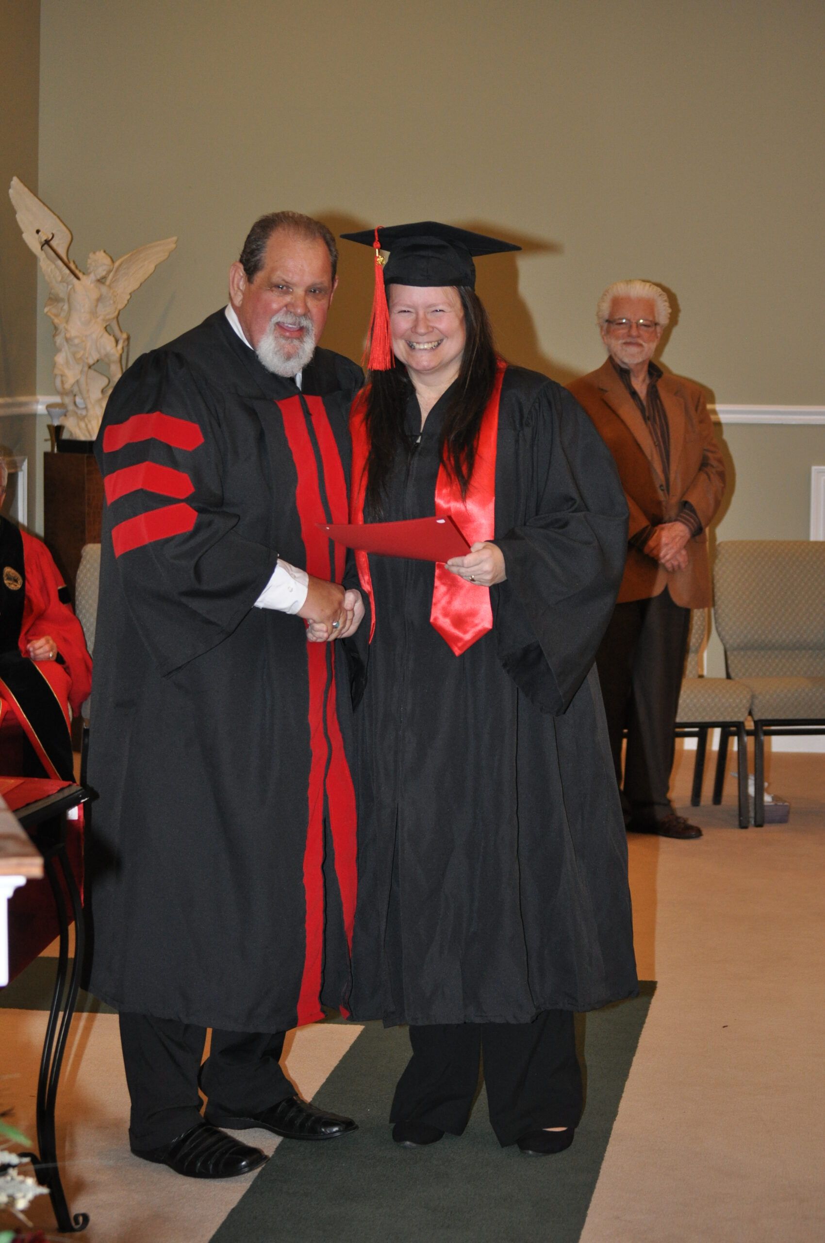 Graduation ceremony: Man in robe hands diploma to person in cap and gown, other man watches.