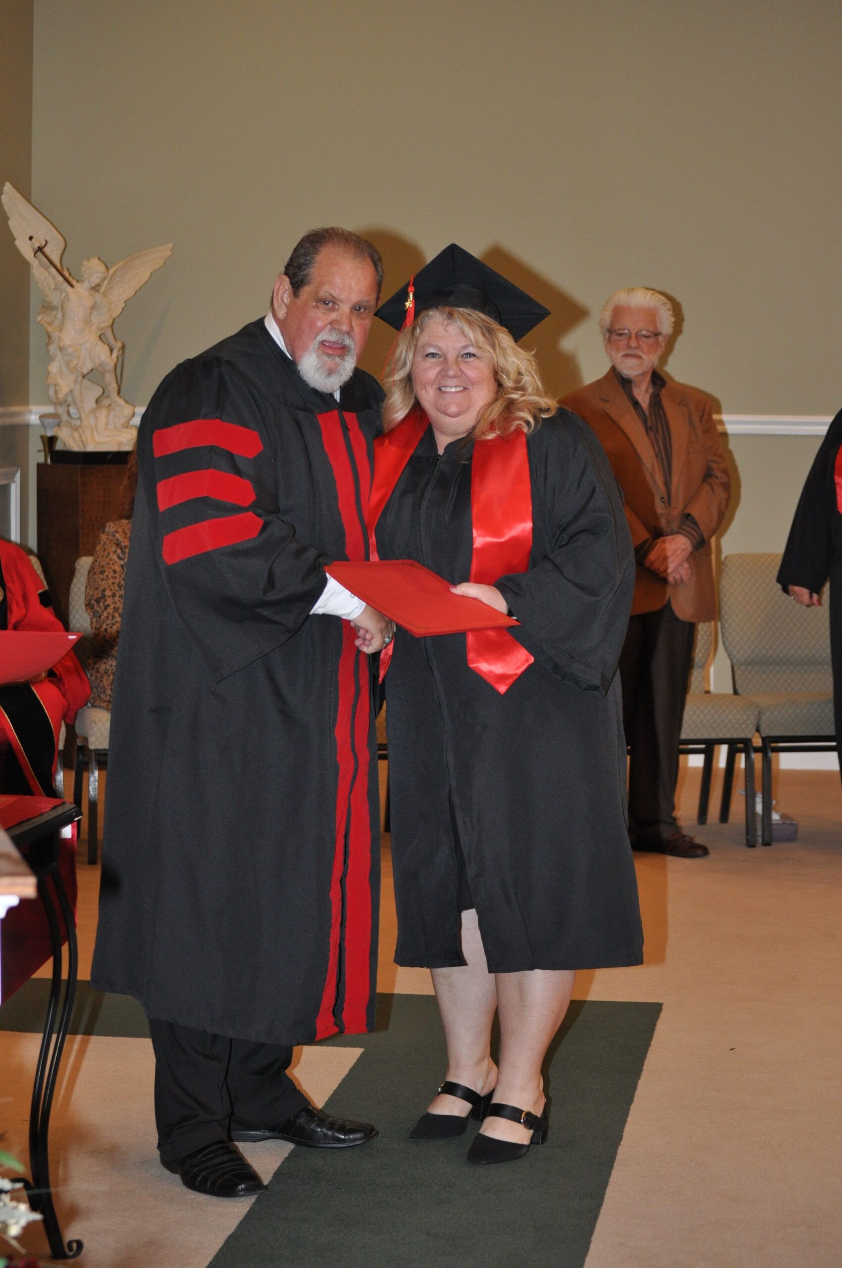 Professor in black and red academic regalia presents a diploma to a smiling graduate in a cap and gown.