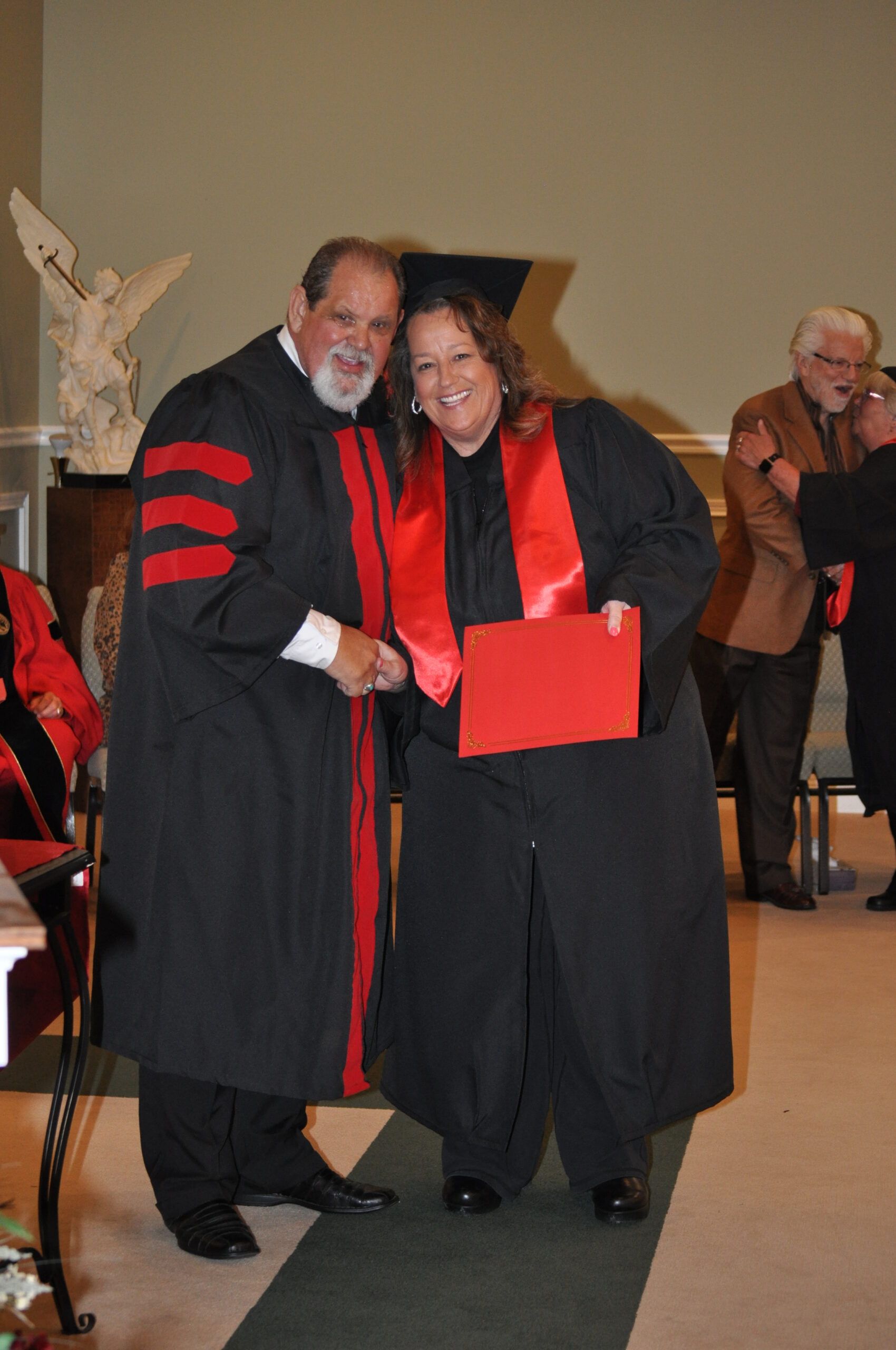 Man in robe presents diploma to woman in robe; both smile. Formal setting with others in the background.