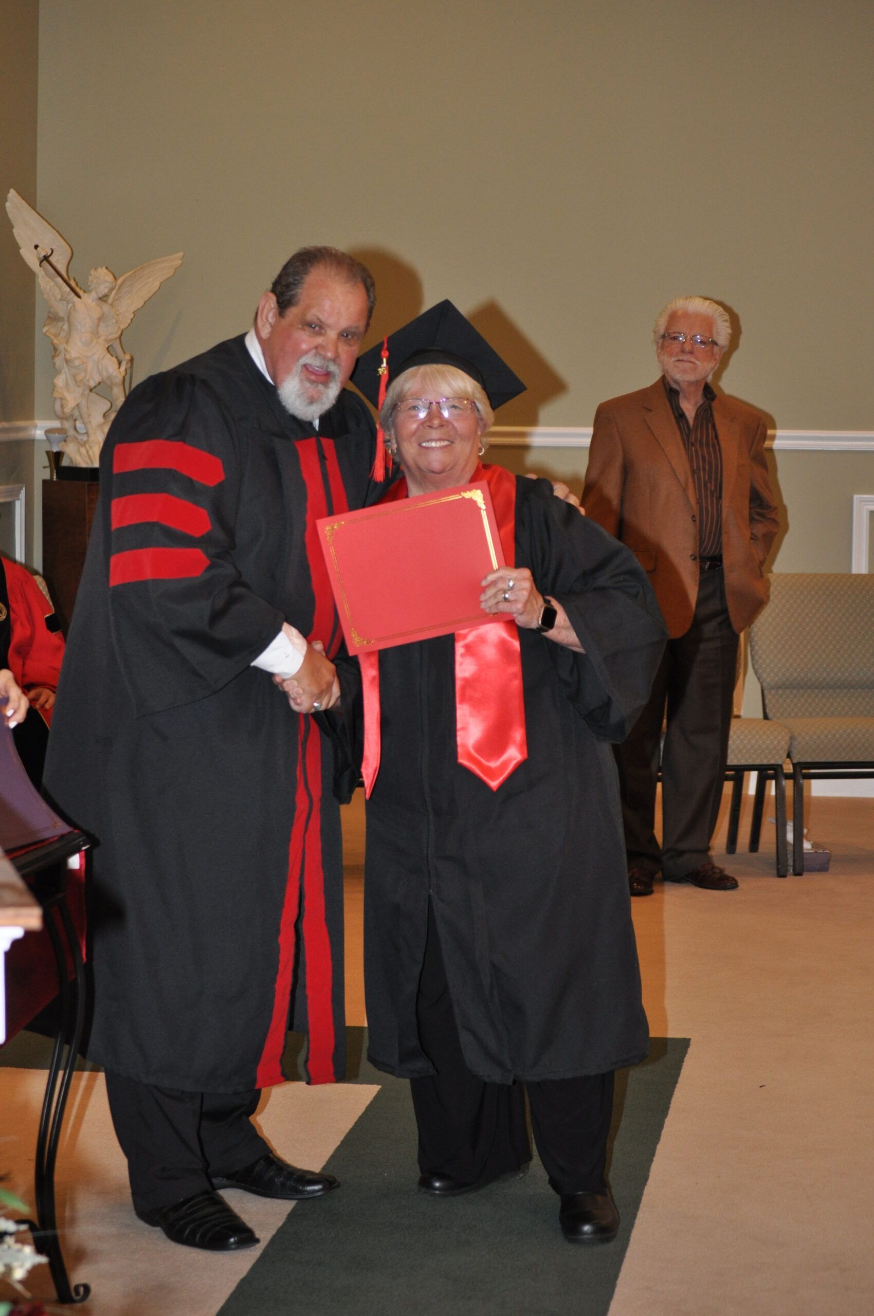 Person in graduation gown receives diploma from robed person, indoors.