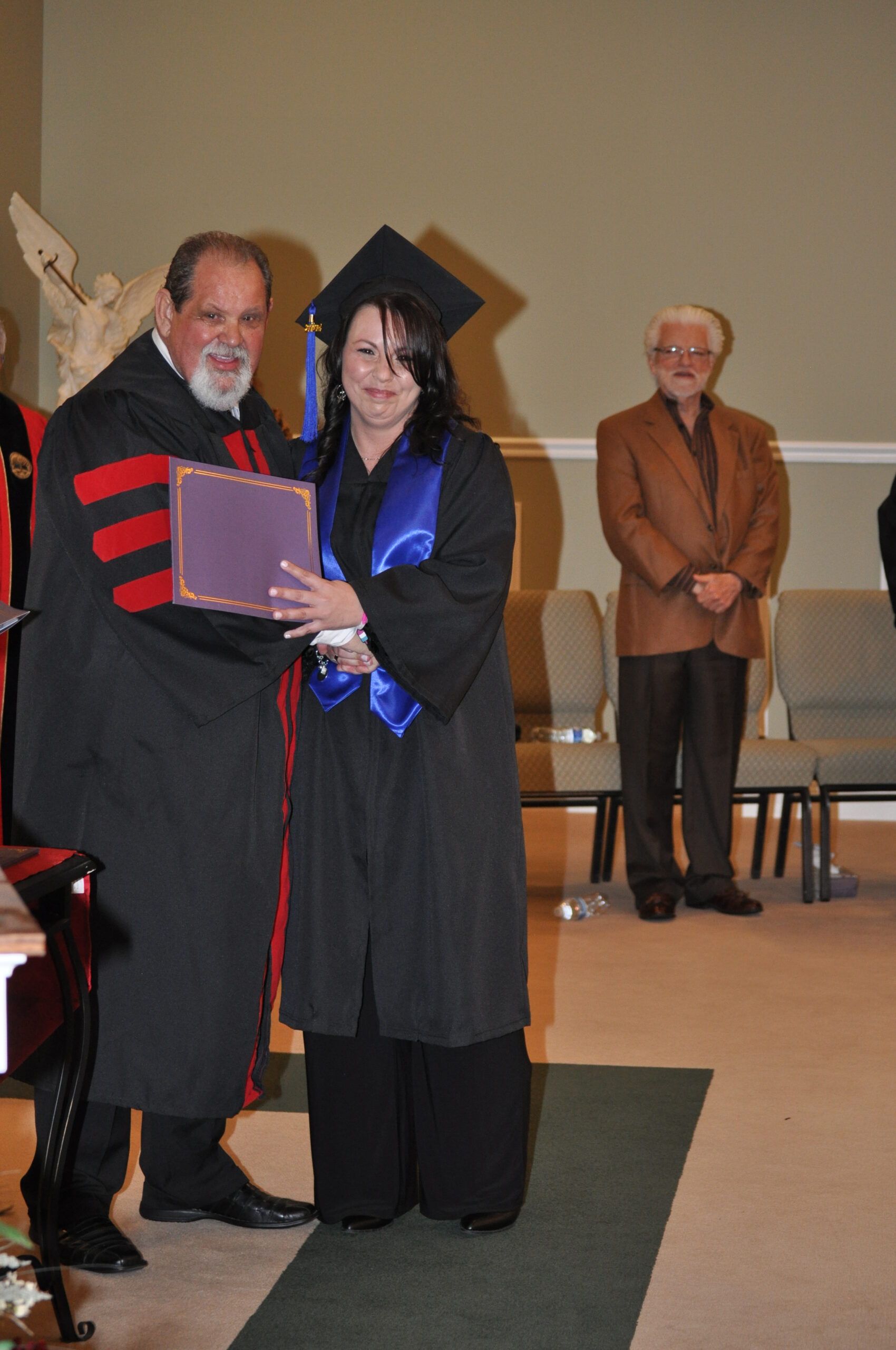 Graduation ceremony: A person in cap and gown receives a diploma from a robed figure. Another person stands in the background.