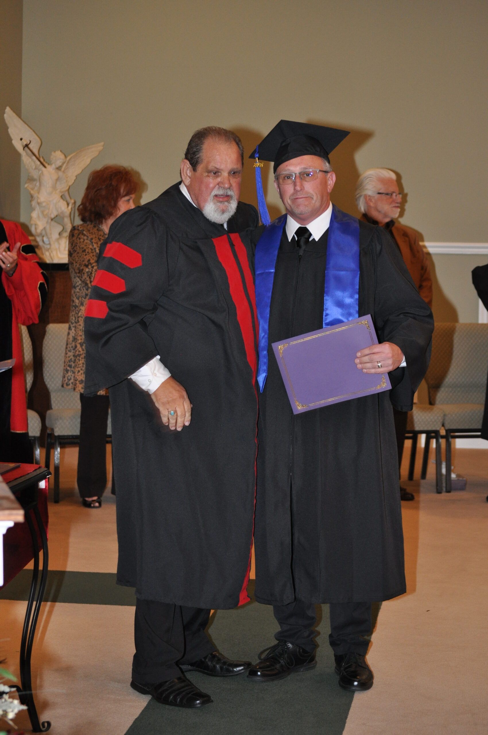 Man in graduation gown receives diploma from robed person, indoors.