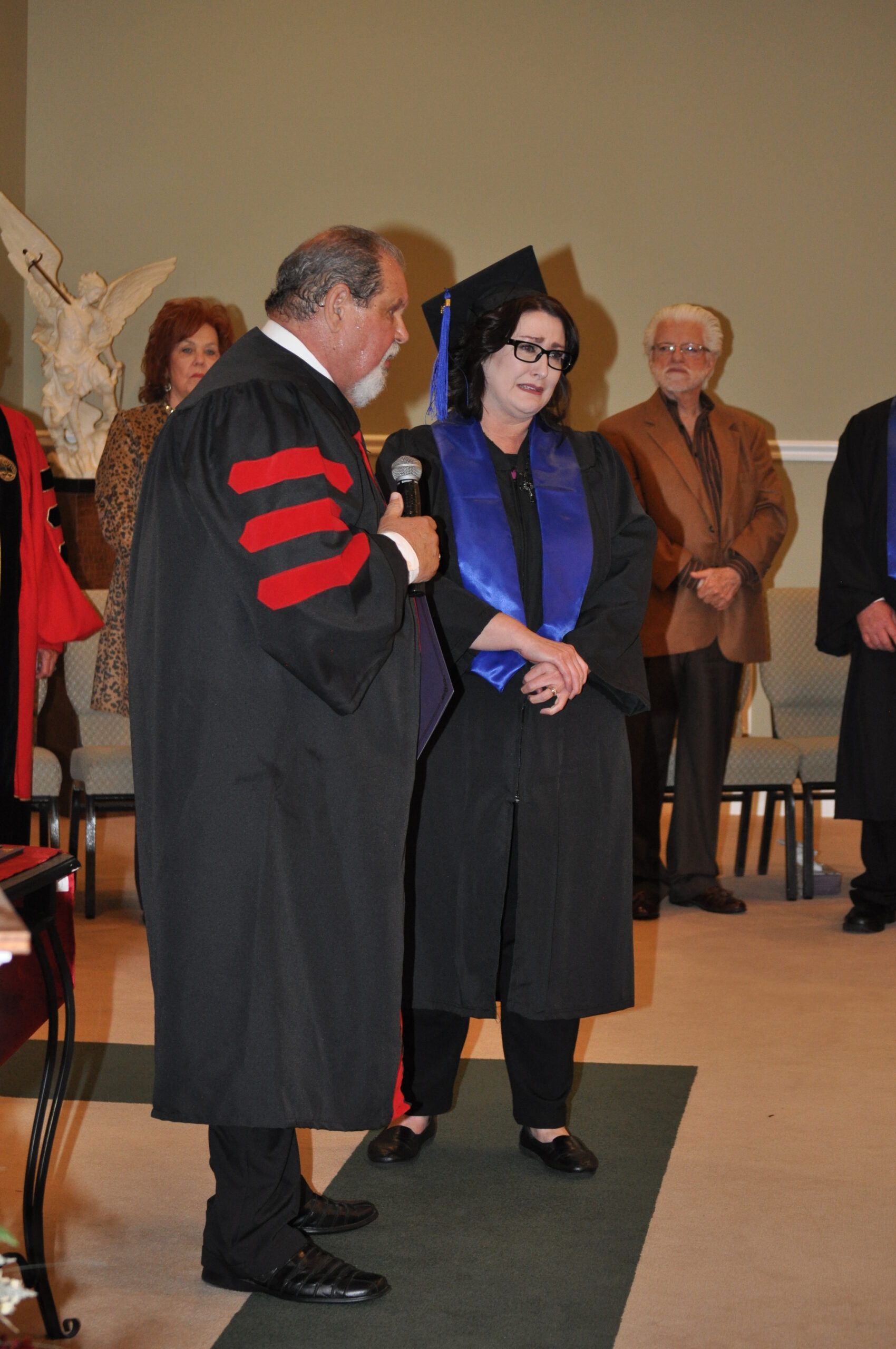 Person in graduation attire being addressed at a ceremony, others watch.