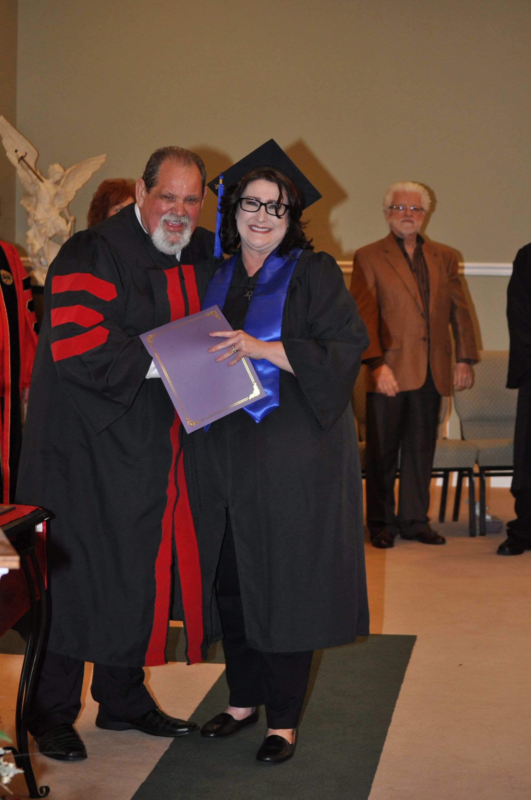 Graduation ceremony: a woman in cap and gown receives a diploma from a robed figure. Others watch.