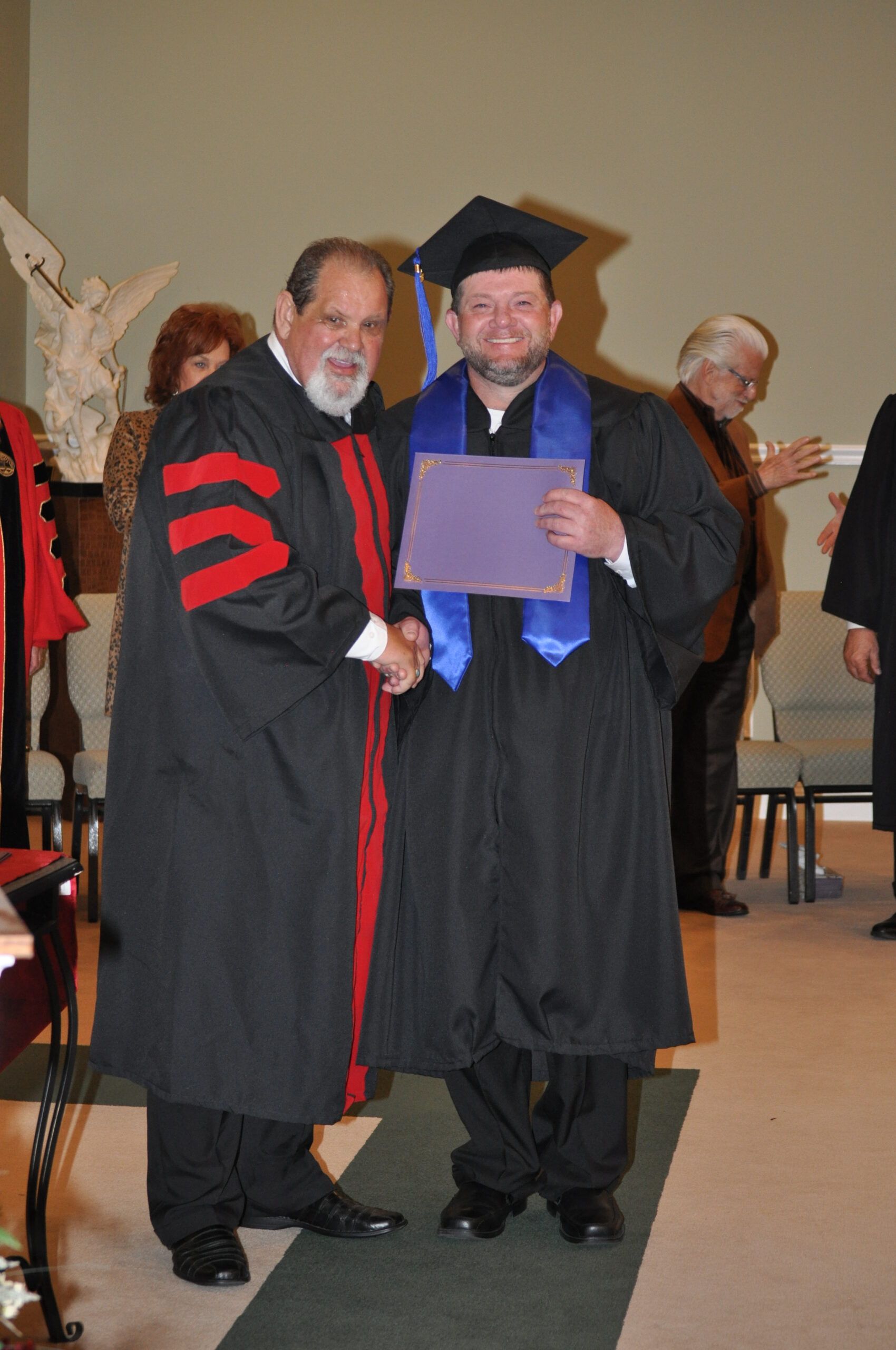 Graduation ceremony: Man in cap and gown shakes hands, receives diploma from robed official.