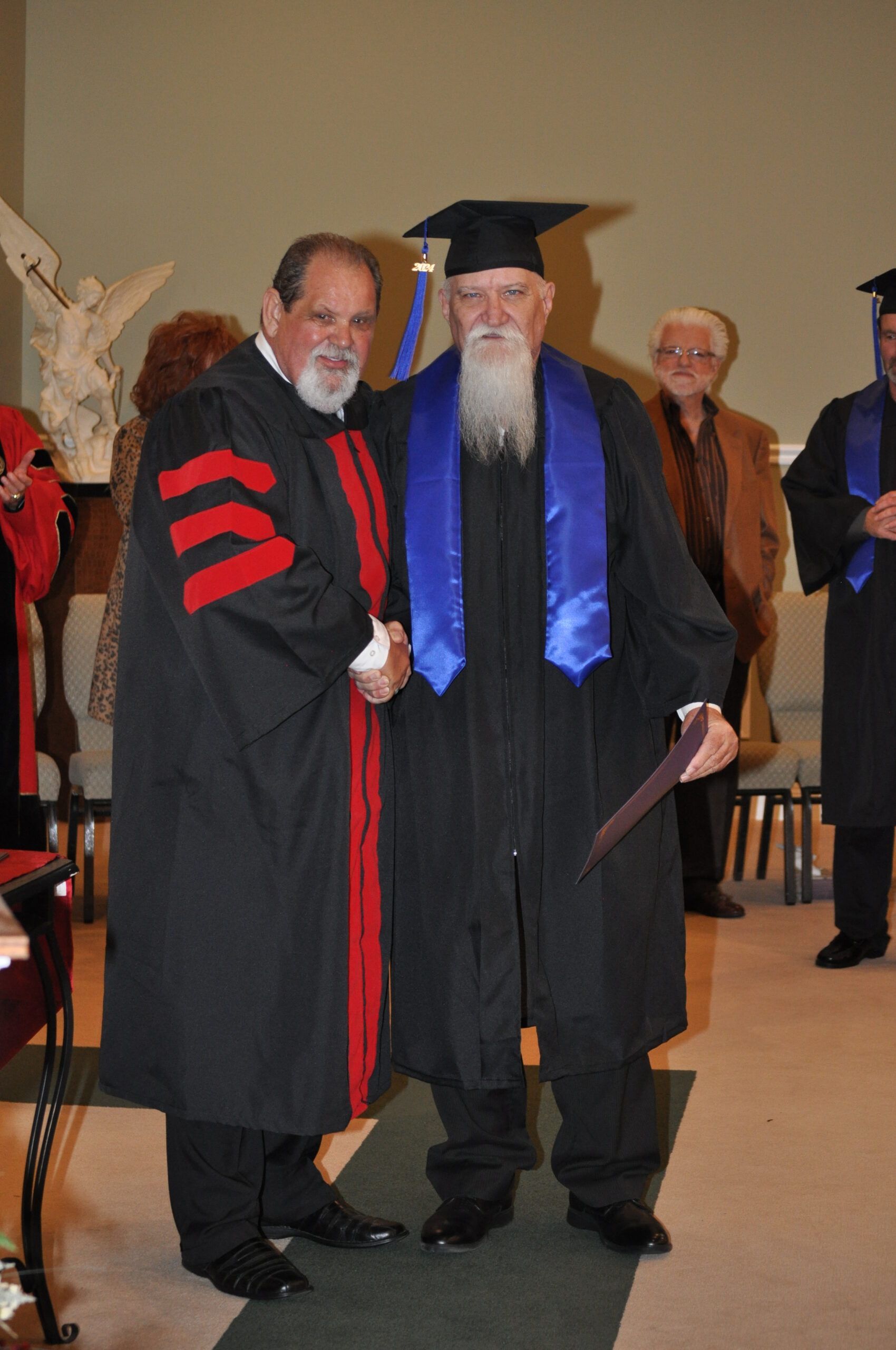 Two men in graduation robes shaking hands, one with a long white beard.