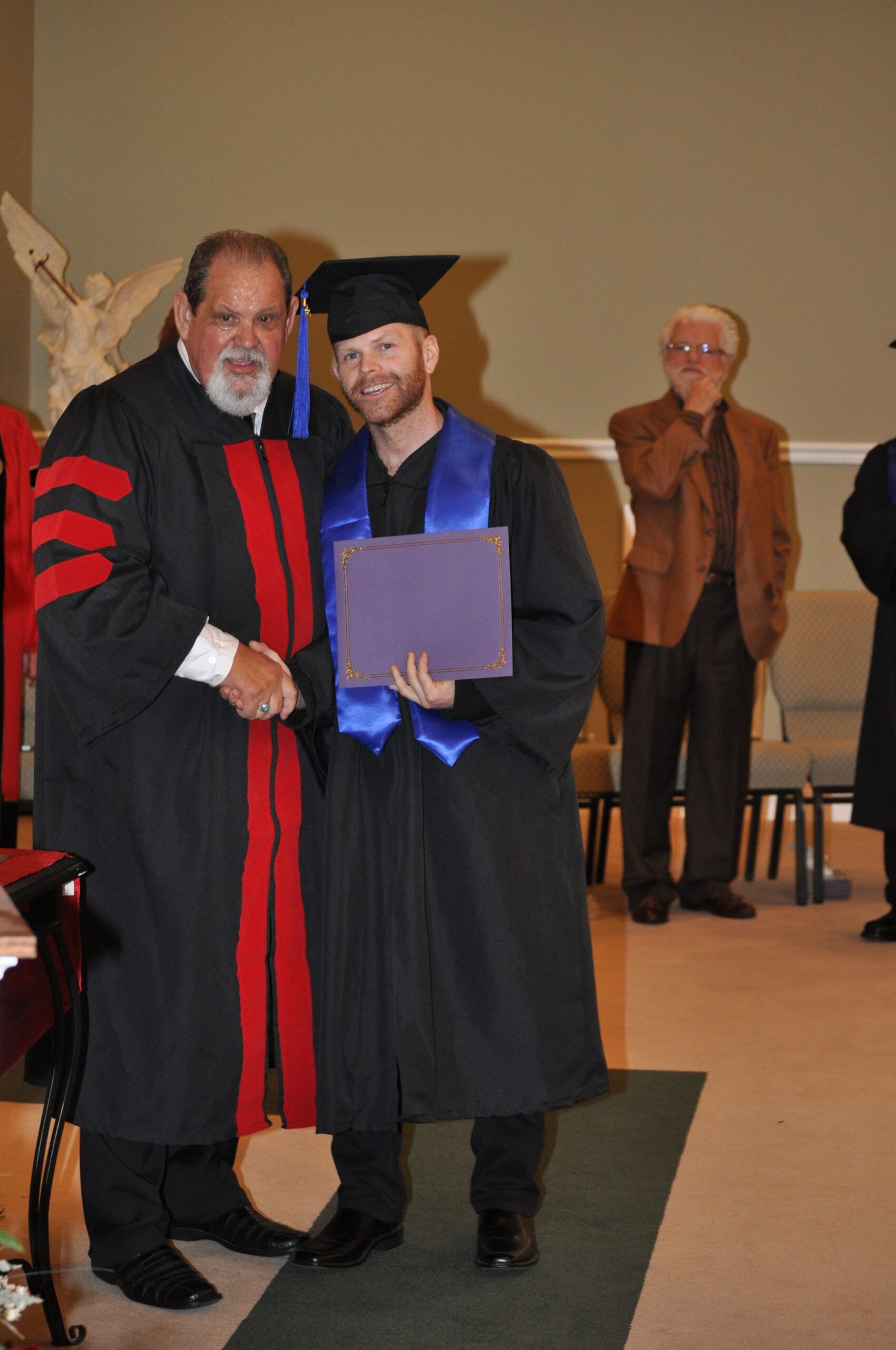 Man in graduation gown shakes hands with a robed figure, holding a purple diploma. A man looks on.