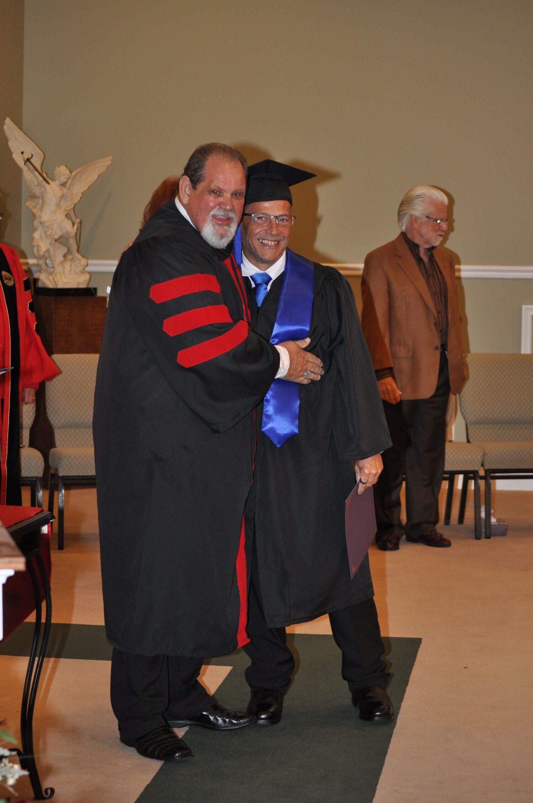 Man in graduation gown being embraced by a robed man, indoors. Both are smiling.