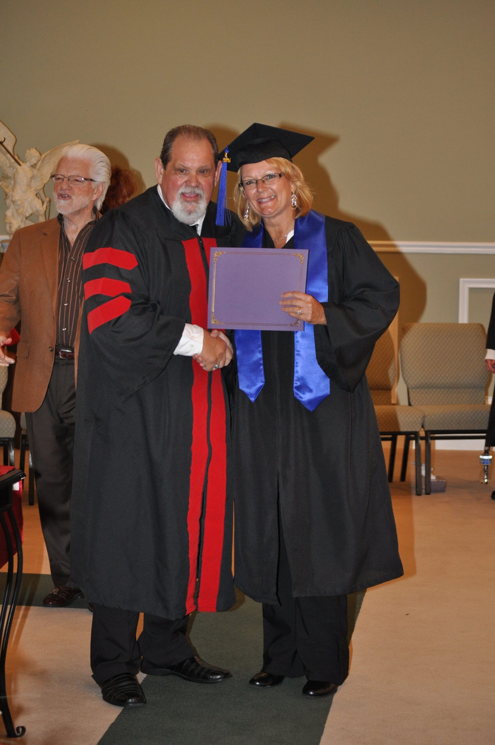 Man in academic robe shakes hands with graduate wearing cap and gown.
