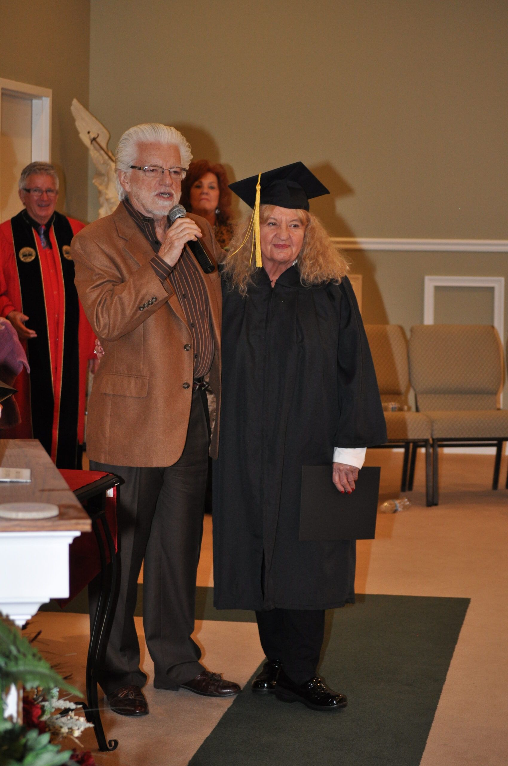 A woman in graduation attire stands beside a man with a microphone in what appears to be a church.