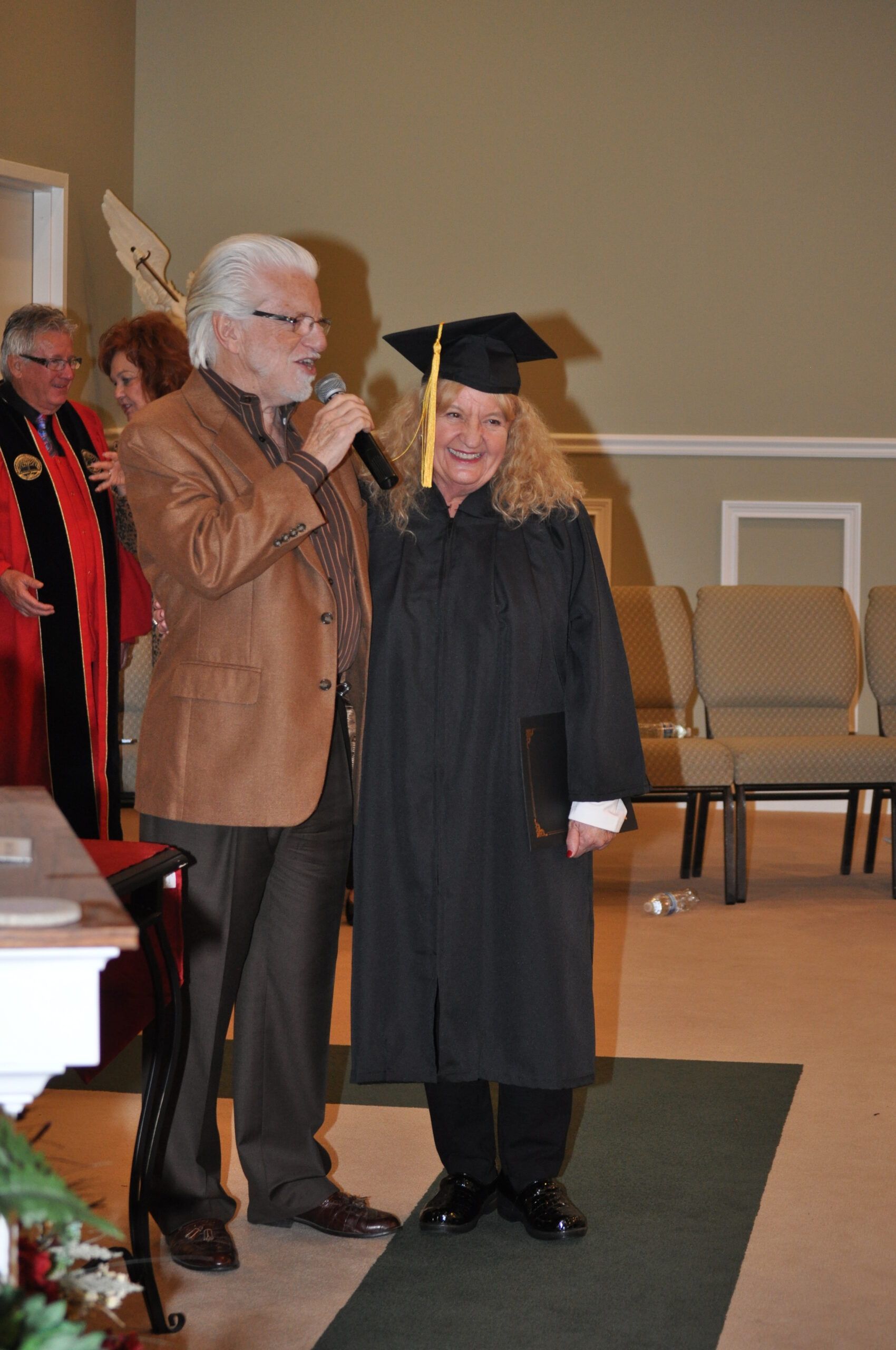Man in brown blazer speaks into microphone next to a person in graduation cap and gown. Inside a building.