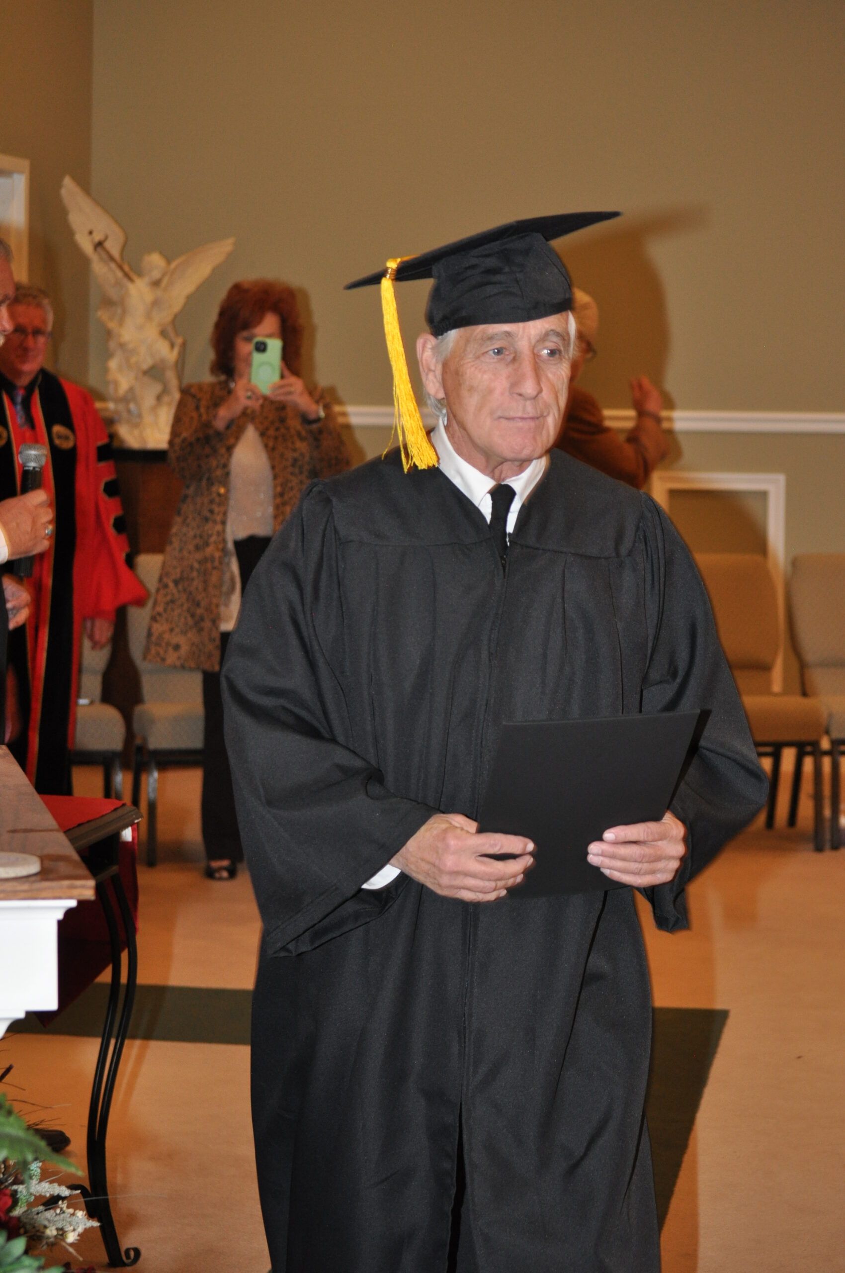 Man in graduation gown and cap holding diploma, indoors.