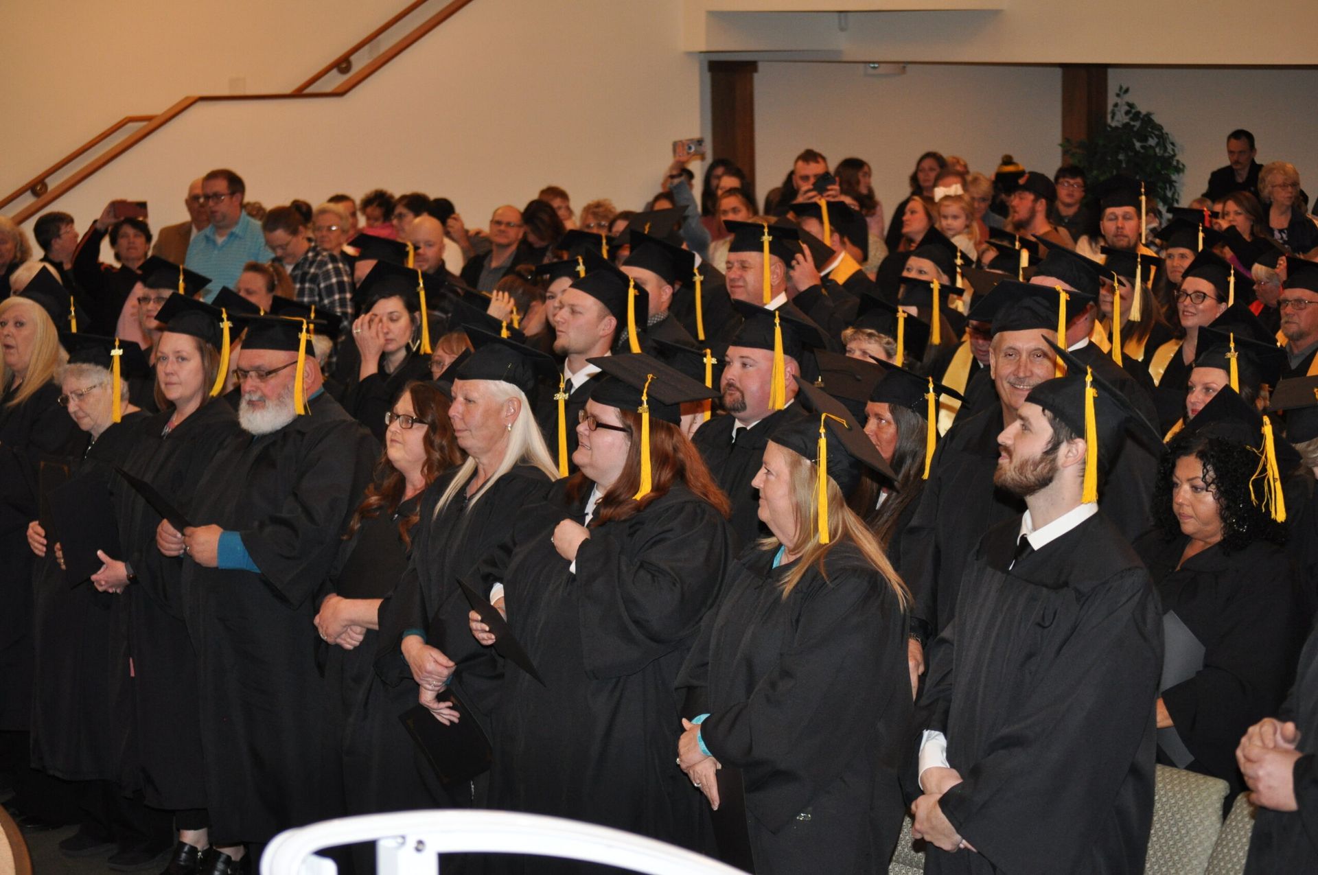 Graduates in black caps and gowns at a ceremony, standing in a crowded room with a wooden-panelled wall.