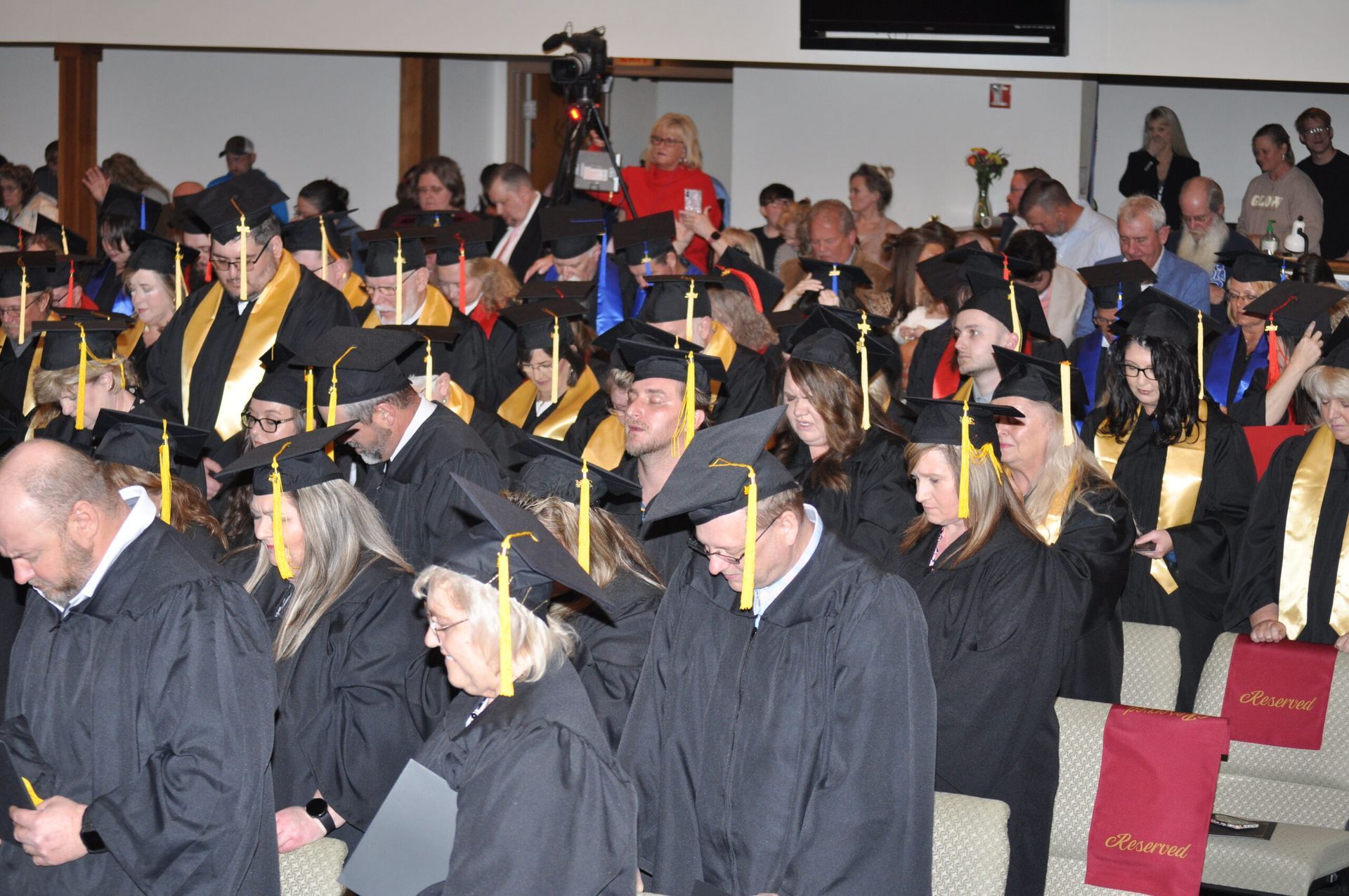 Graduation ceremony: graduates in black gowns, mortarboards, gold stoles, seated indoors, faces down.