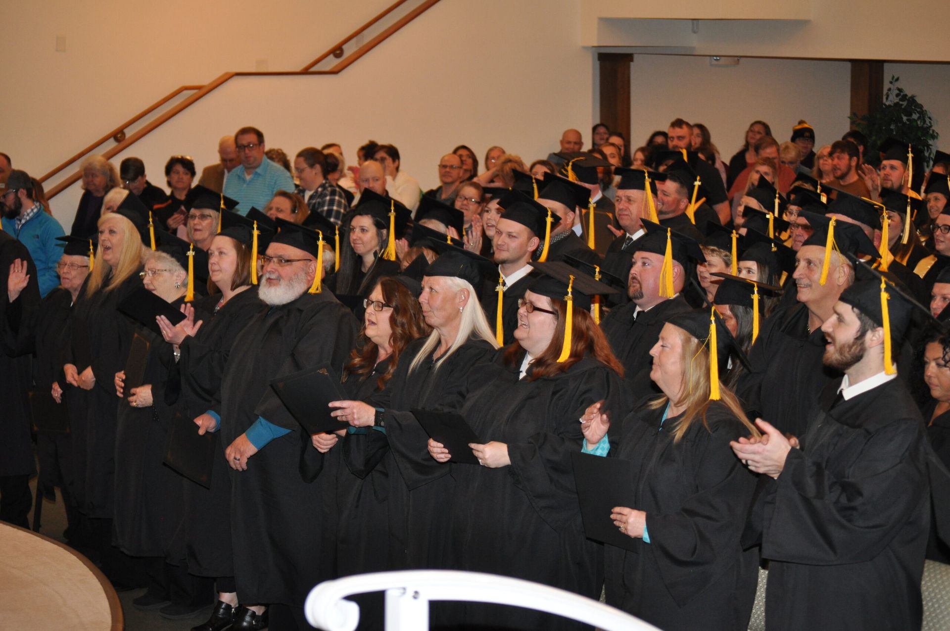 Graduation ceremony: graduates in black gowns and caps, some clapping, in an auditorium.